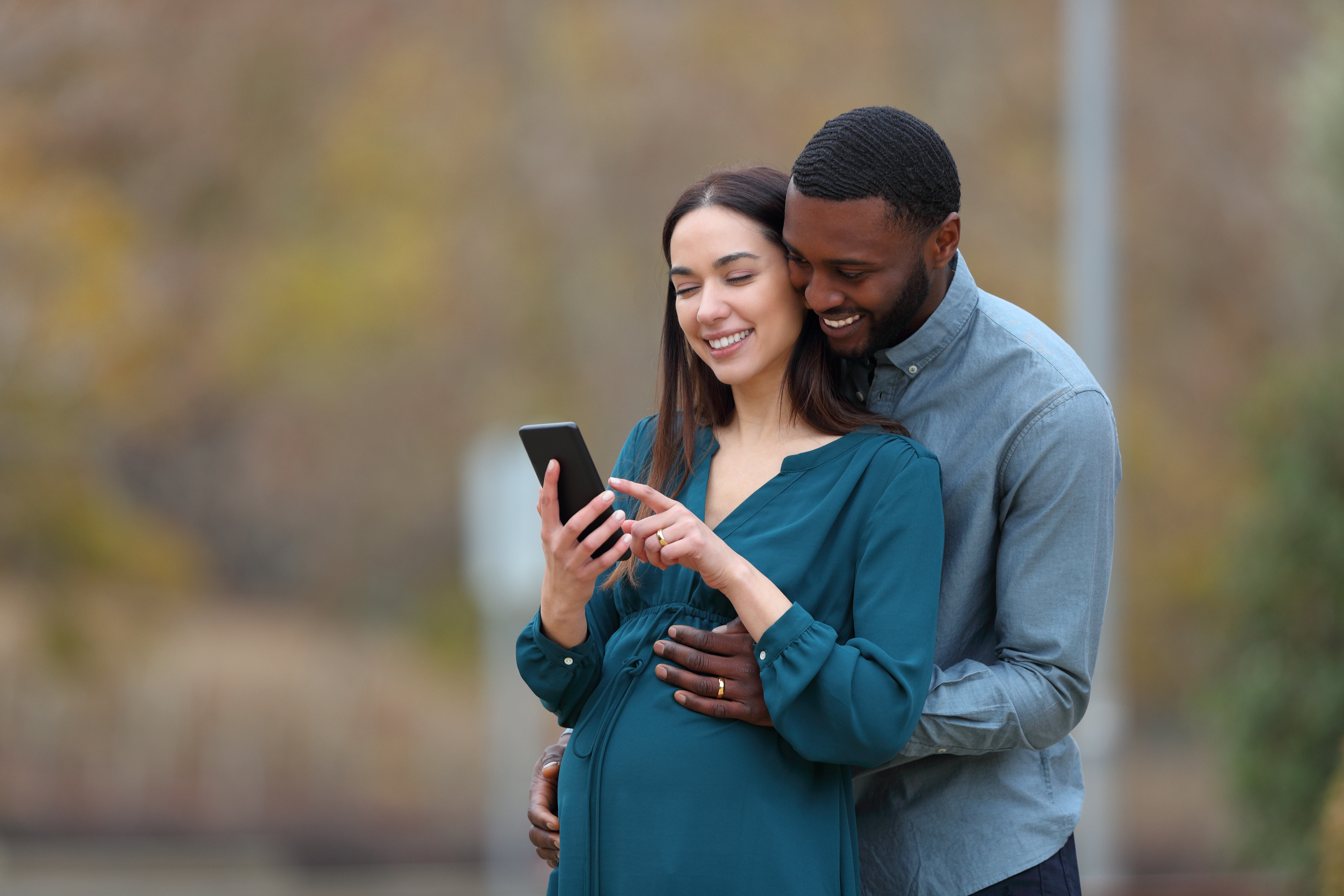 An interracial couple with a pregnant wife and her husband checking smartphone in a park | Source: Shutterstock