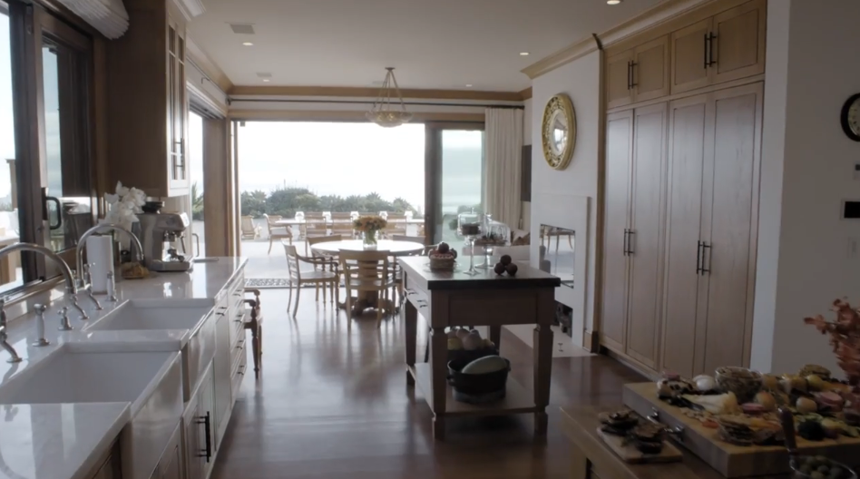Looking through the expansive open-concept kitchen, polished wooden floors reflect light streaming in from the large oceanfront glass doors at the far end. A central prep island sits between built-in light wood pantry cabinetry and double farm sinks, leading seamlessly toward the sunlit breakfast nook overlooking the terrace. | Source: YouTube/Architectural Digest