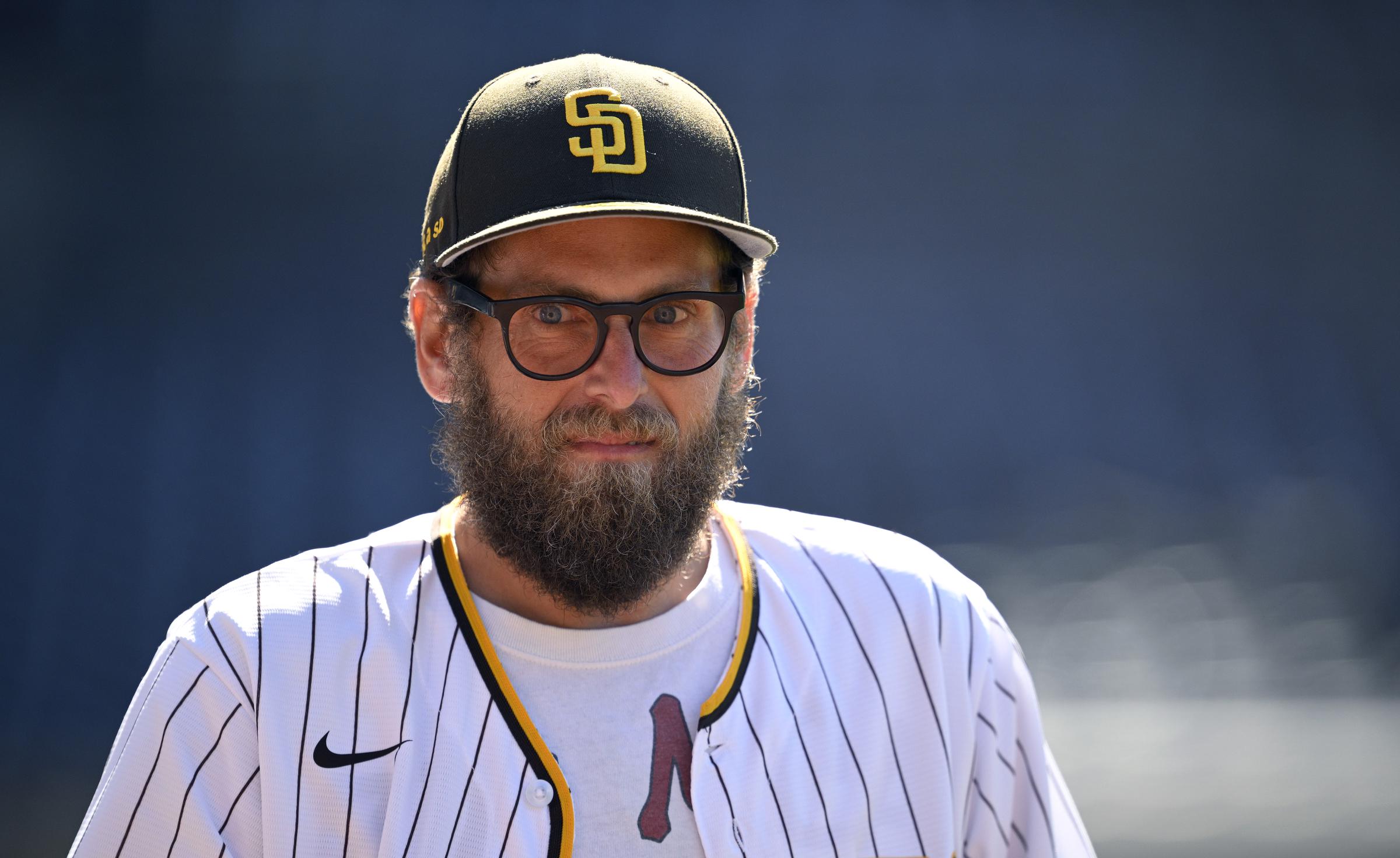 Jonah Hill is seen at a San Diego Padres game wearing the team’s pinstriped jersey and a matching cap. With a full beard, round glasses, and a serious expression, he stands under the sun on the field.