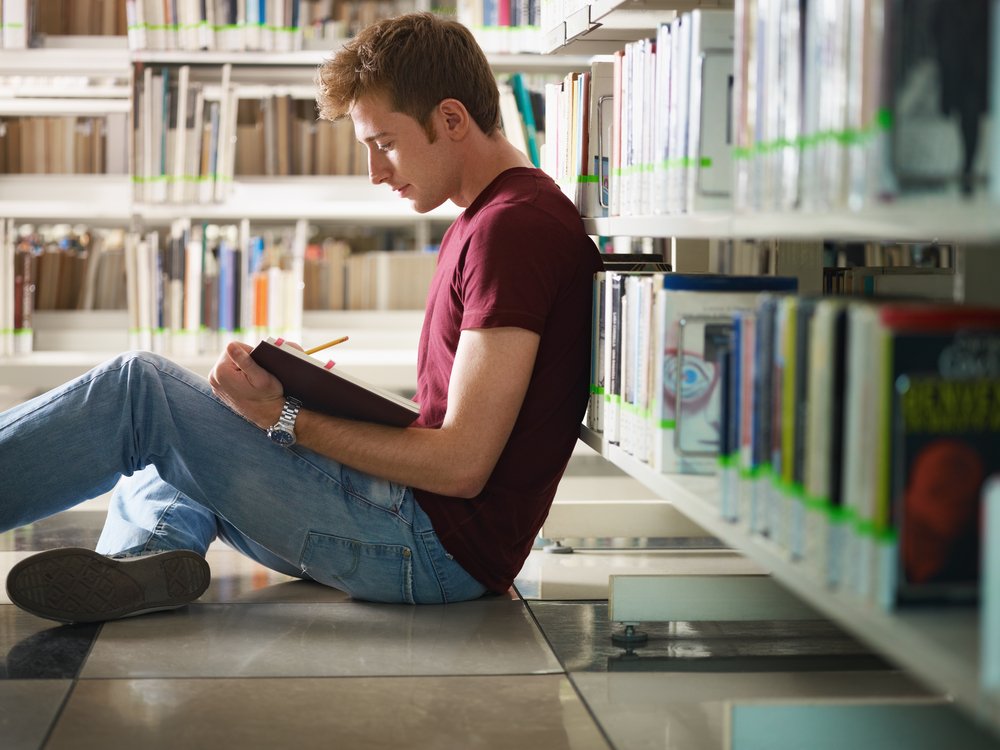Joven sentado en el suelo del pasillo de una biblioteca estudiando. | Foto: Shutterstock