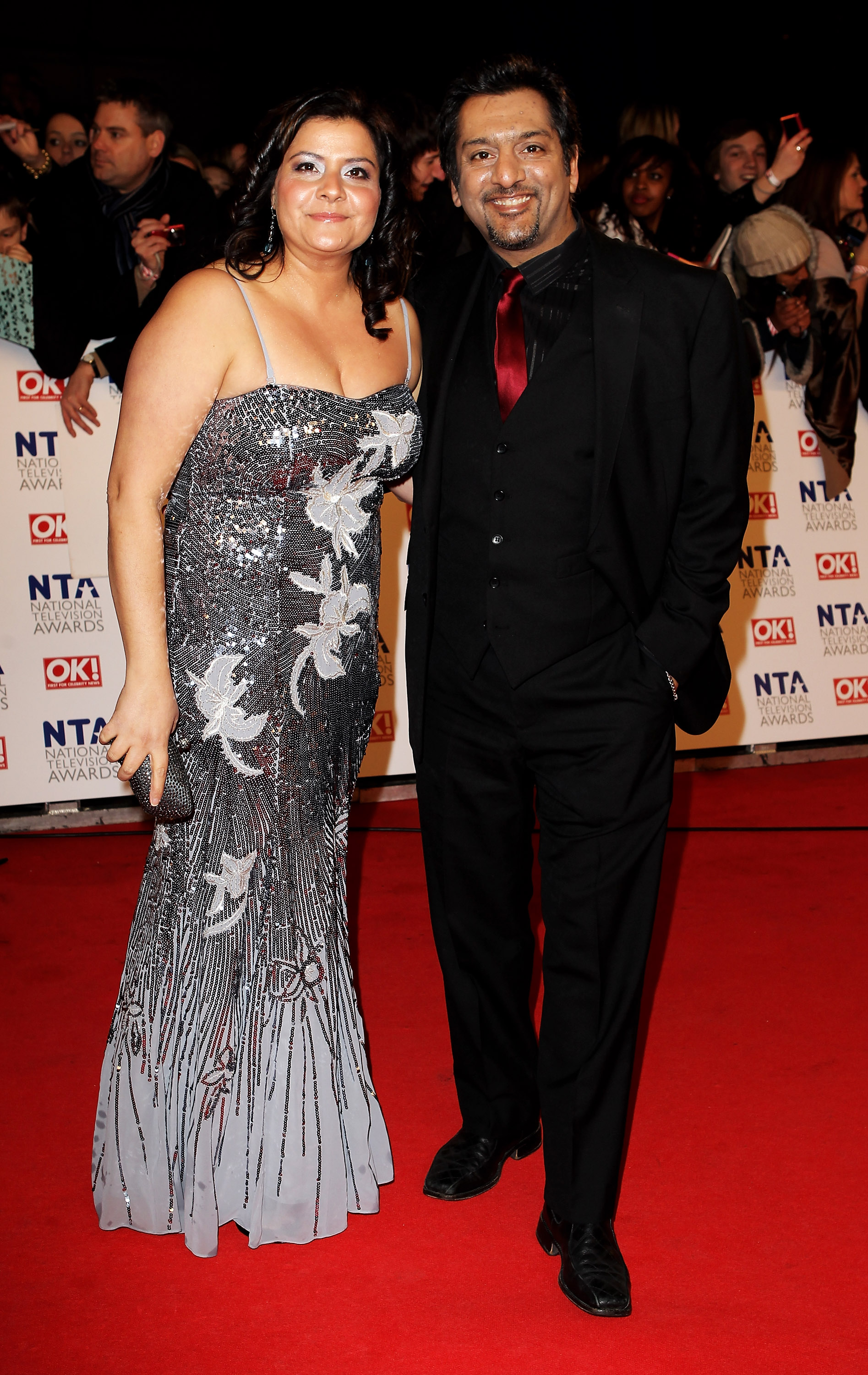 Wadia poses alongside actor Nitin Ganatra on the red carpet at the National Television Awards at the O2 Arena in January 2011. She wears a striking floor-length silver beaded gown with large white floral appliqué detail and a fitted mermaid silhouette, while Ganatra stands beside her in an all-black suit with a deep red tie.