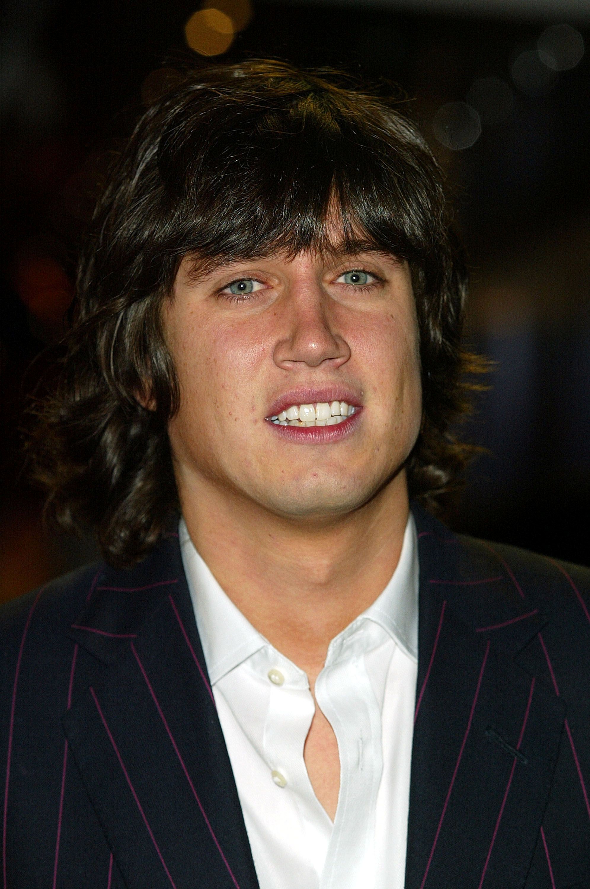 Vernon Kay wears a dark pinstriped blazer over an open-collar white shirt, offering a relaxed smile as he looks toward the camera.