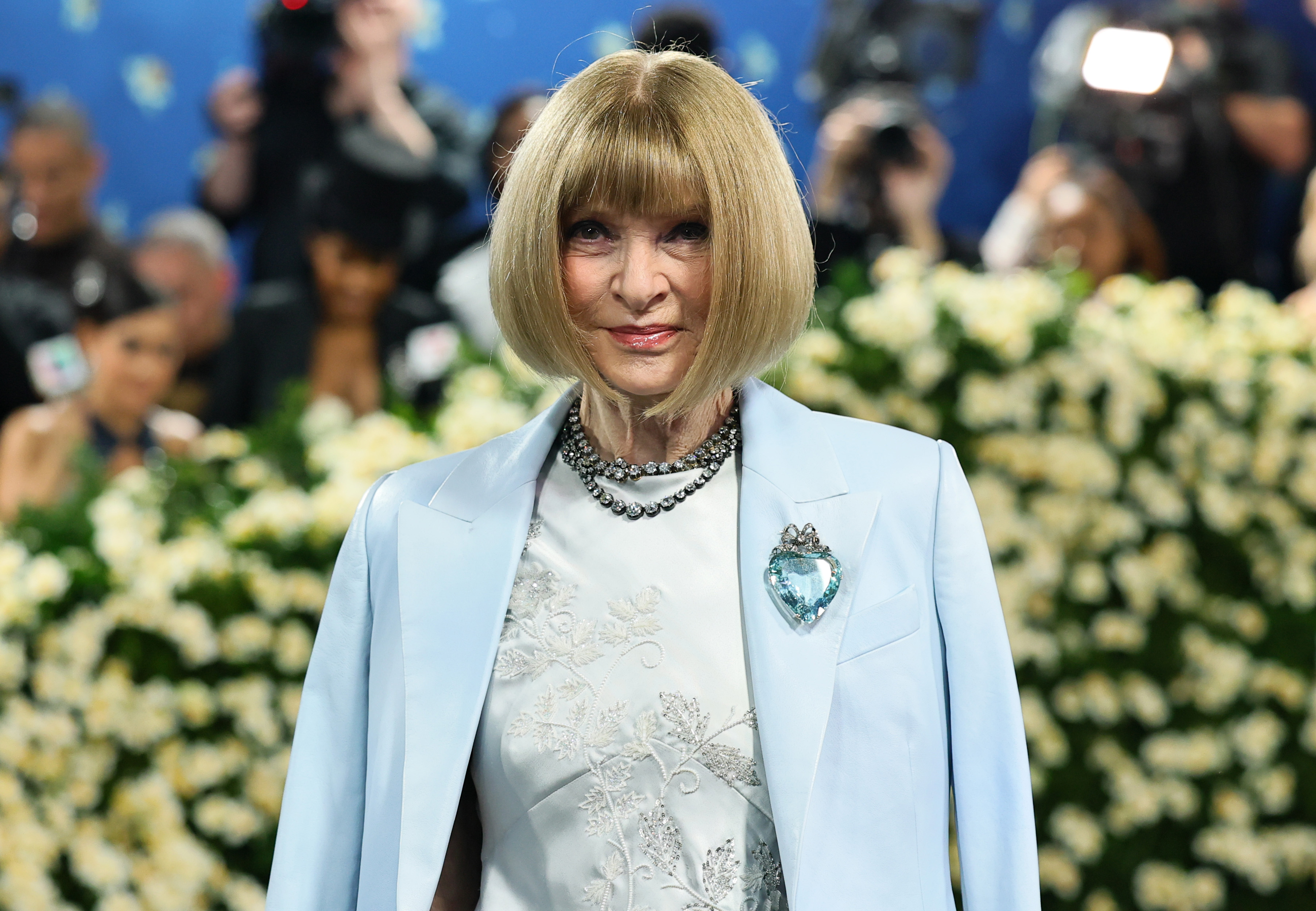 Against a backdrop of white florals, Anna Wintour smiles in her custom ensemble at the 2025 Met Gala. Her look features a silver-embroidered blue dress and glistening necklace for the "Superfine: Tailoring Black Style" theme.