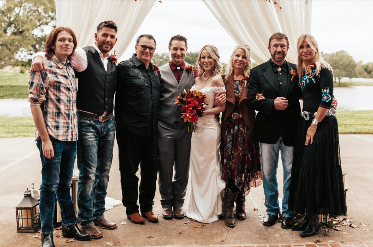 A wedding party gathers beneath a soft, draped arch, with Chuck Norris, Gena O'Kelley Norris, and Dina Norris DiCiolli standing among family and friends. | Source: Facebook/GabiBoardman