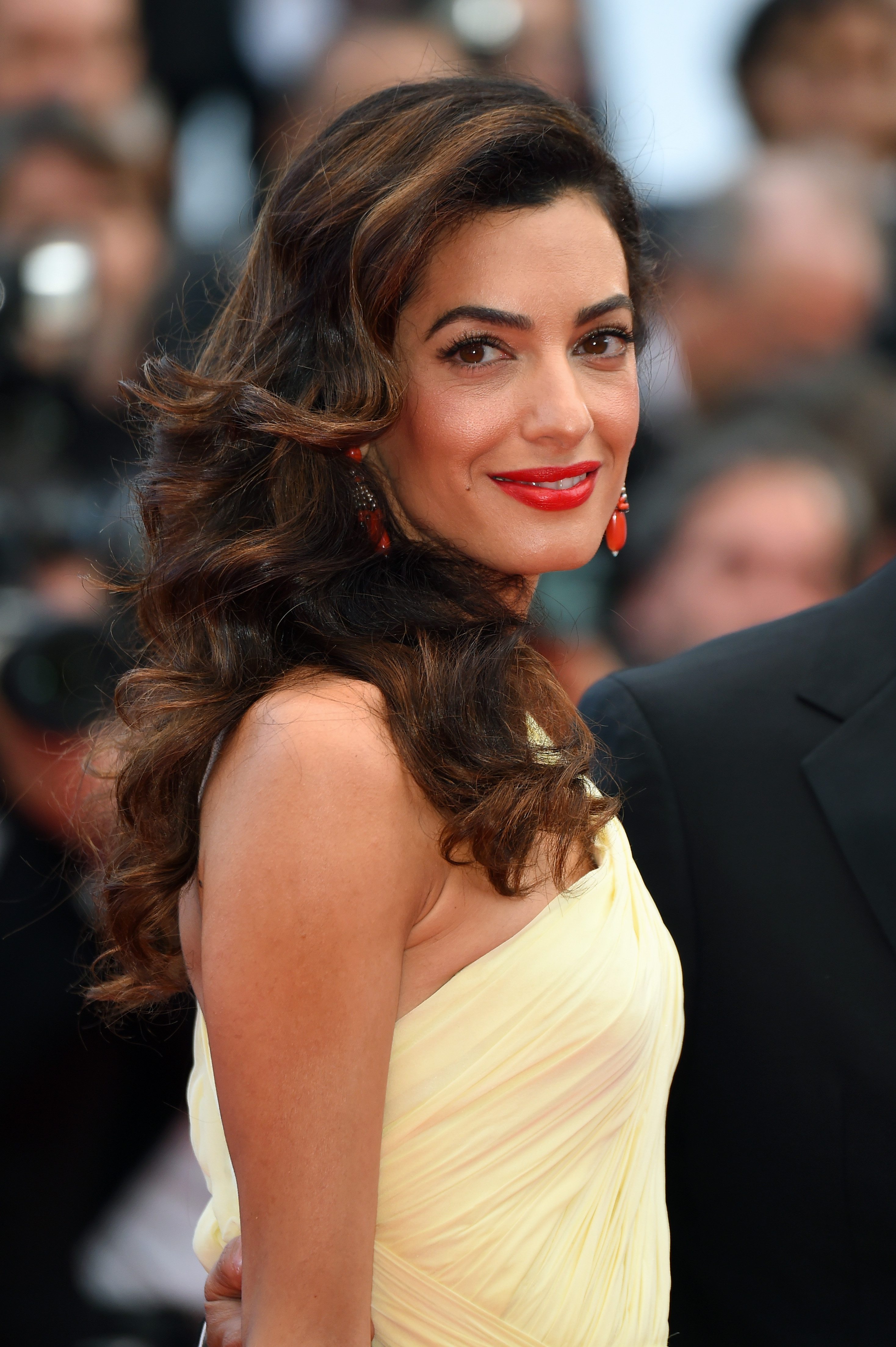 On the red carpet at the Cannes premiere of "Money Monster," Amal stuns in a soft yellow one-shoulder gown with ruched detailing, paired with red drop earrings, a bold red lip, and side-swept voluminous curls that recall vintage Hollywood glamour.