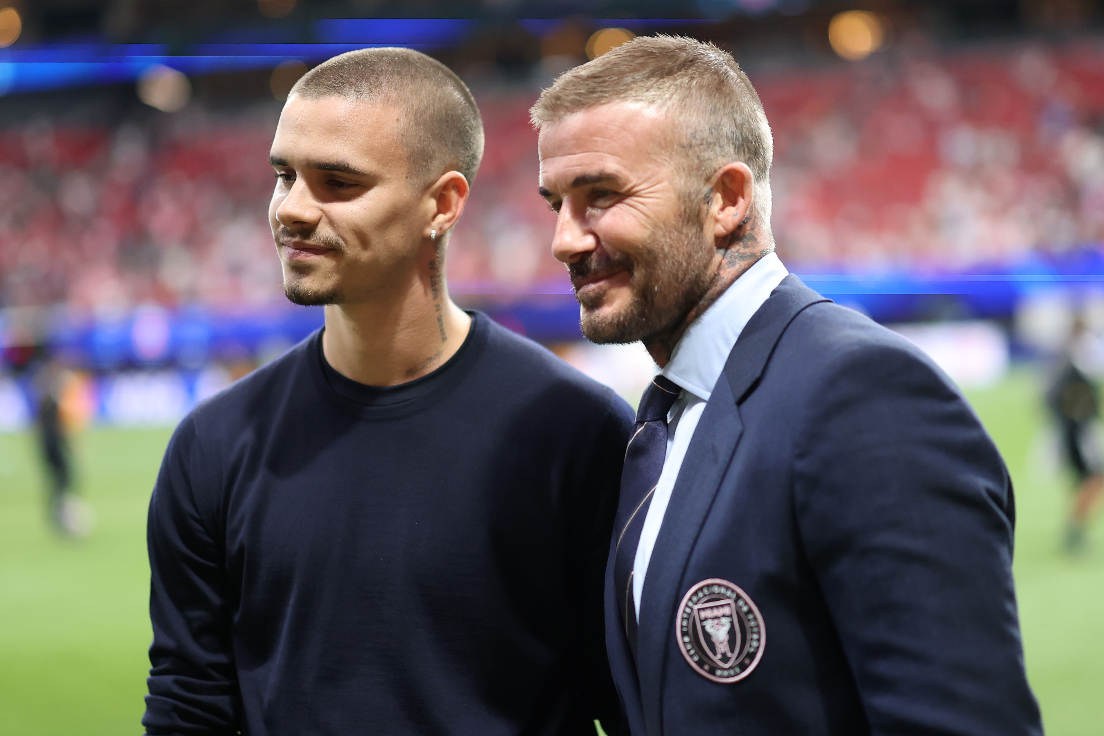 Romeo and David Beckham photographed together pitch-side at the FIFA Club World Cup in Atlanta, June 2025 — Romeo in a navy jumper, David in an Inter Miami blazer, both with the same jaw, ears, and easy, knowing half-smile.
