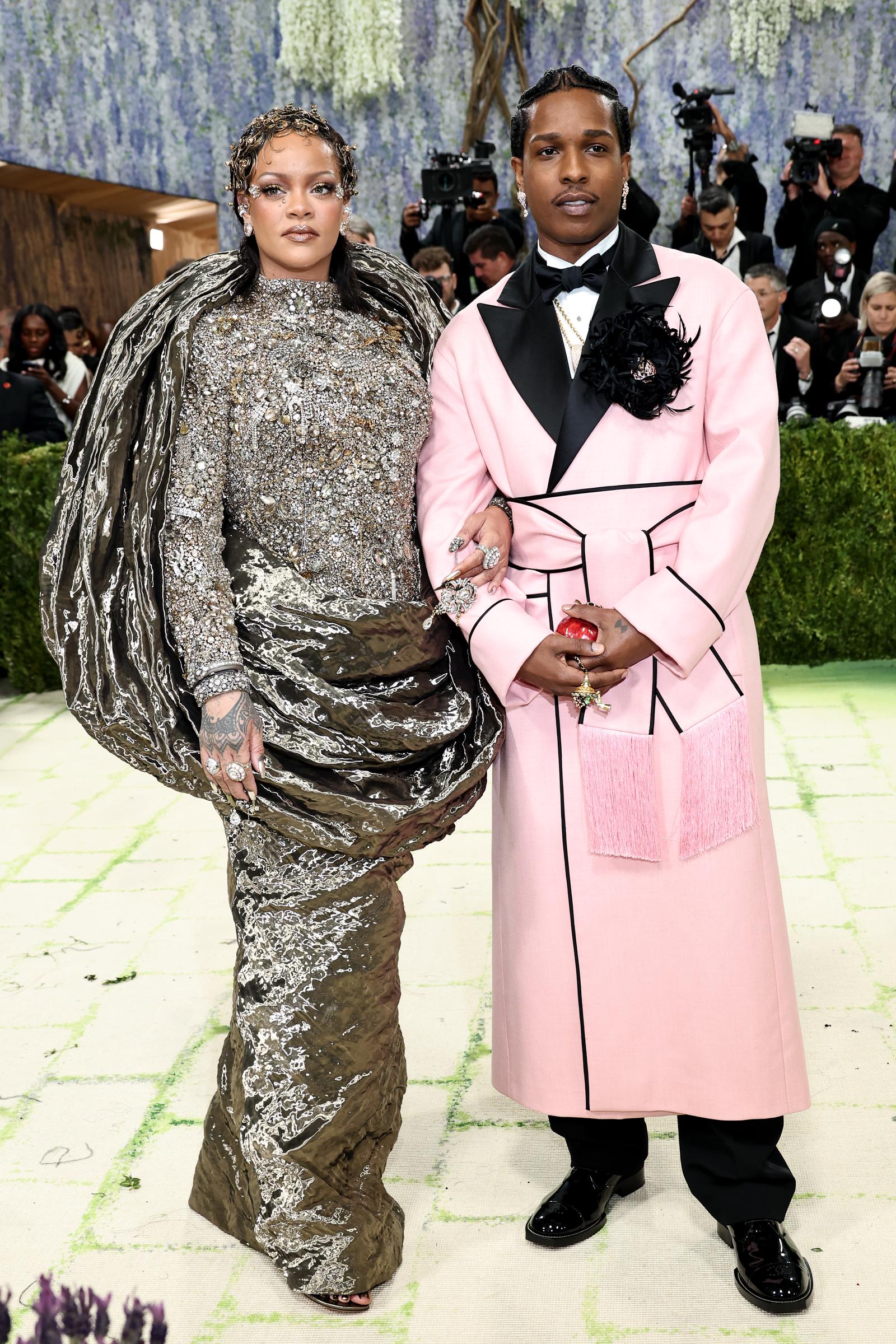 Rihanna makes a statement in a sculptural, jewel-encrusted gown with dramatic metallic draping. A$AP Rocky stands beside her in a structured pink coat with black lapels and floral detailing.