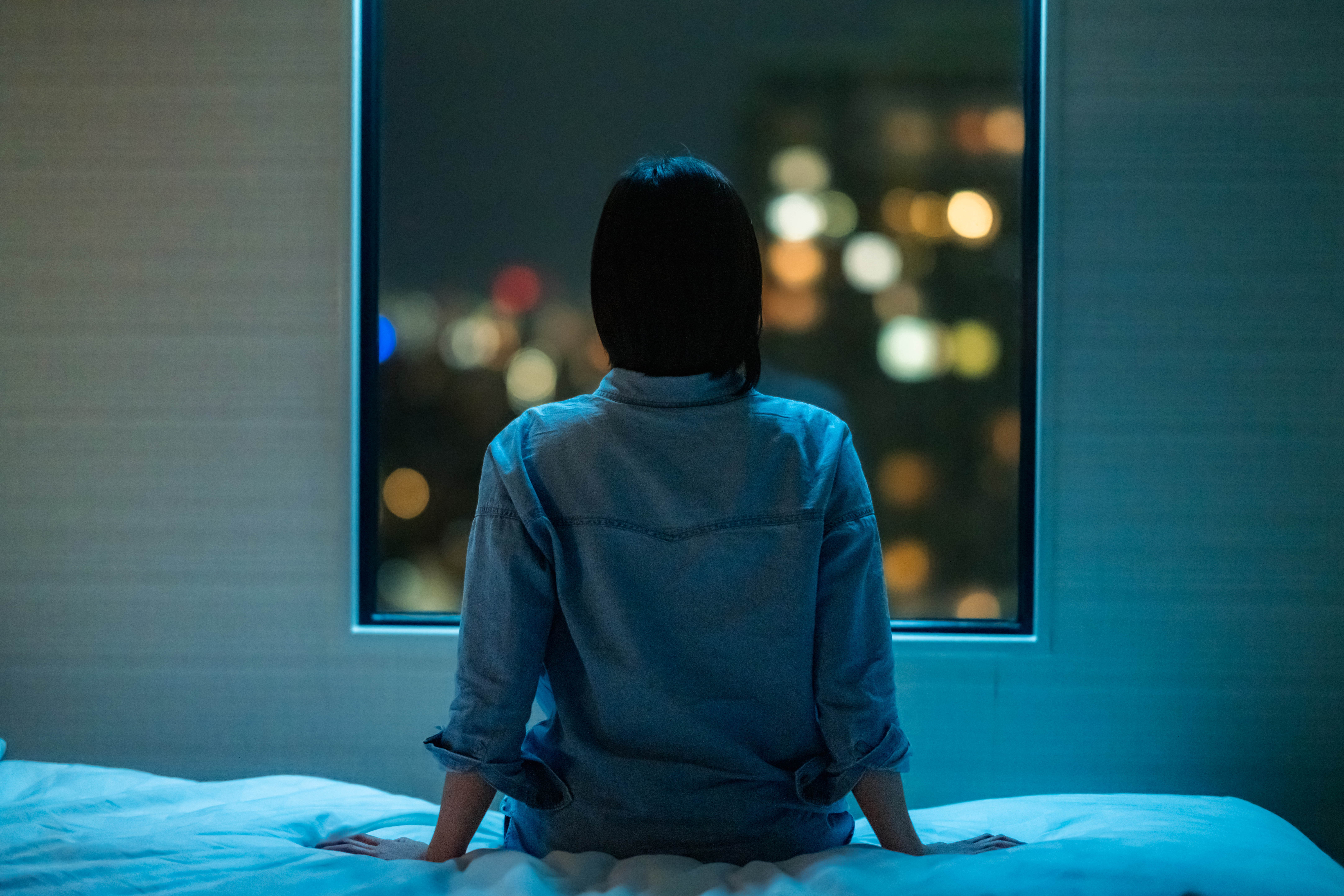 Rear view of woman sitting alone on bed in room and looking through window at night | Source: Getty Images