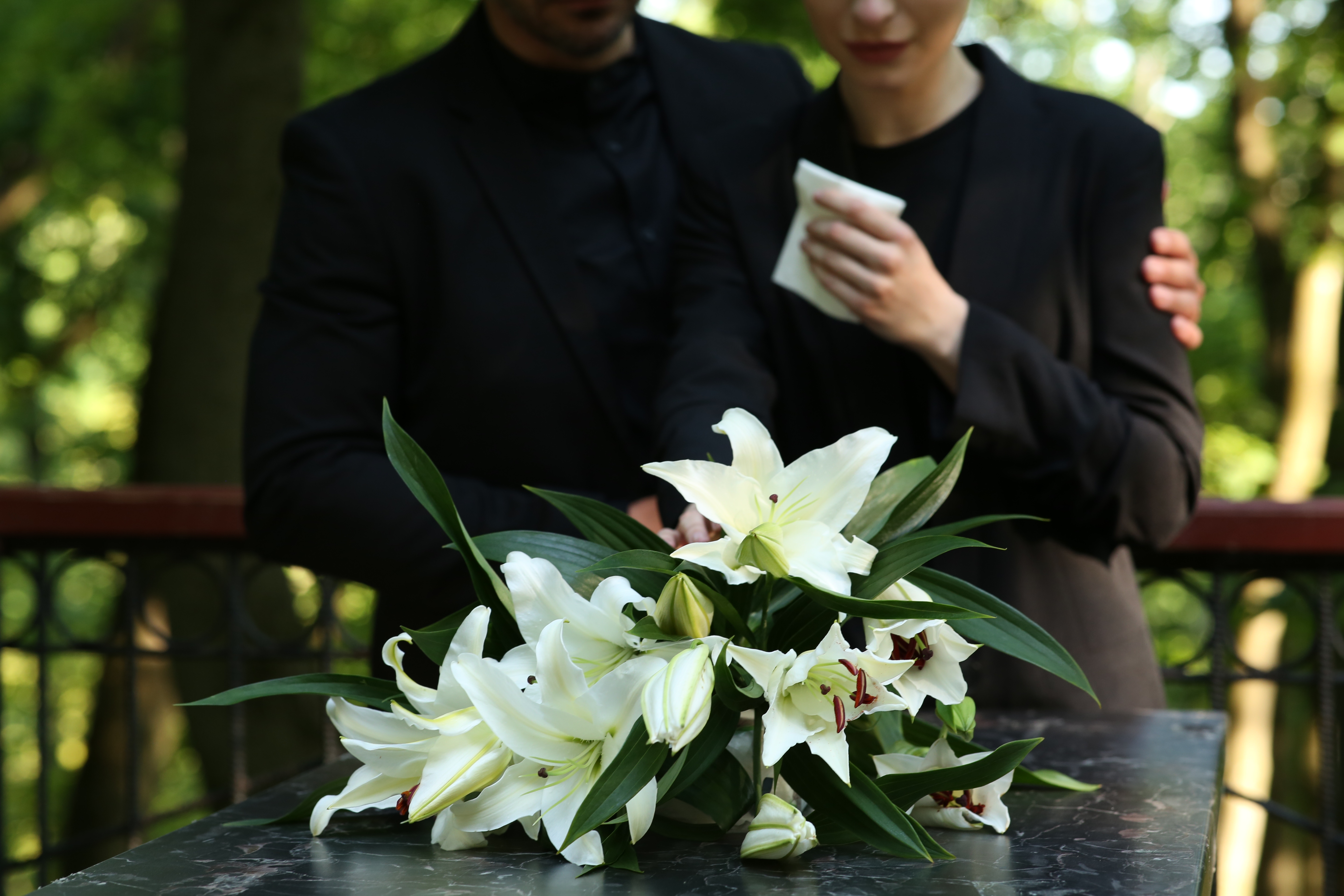 Couple near granite tombstone with white lilies at cemetery outdoors, selective focus. Funeral ceremony | Source: Getty Images