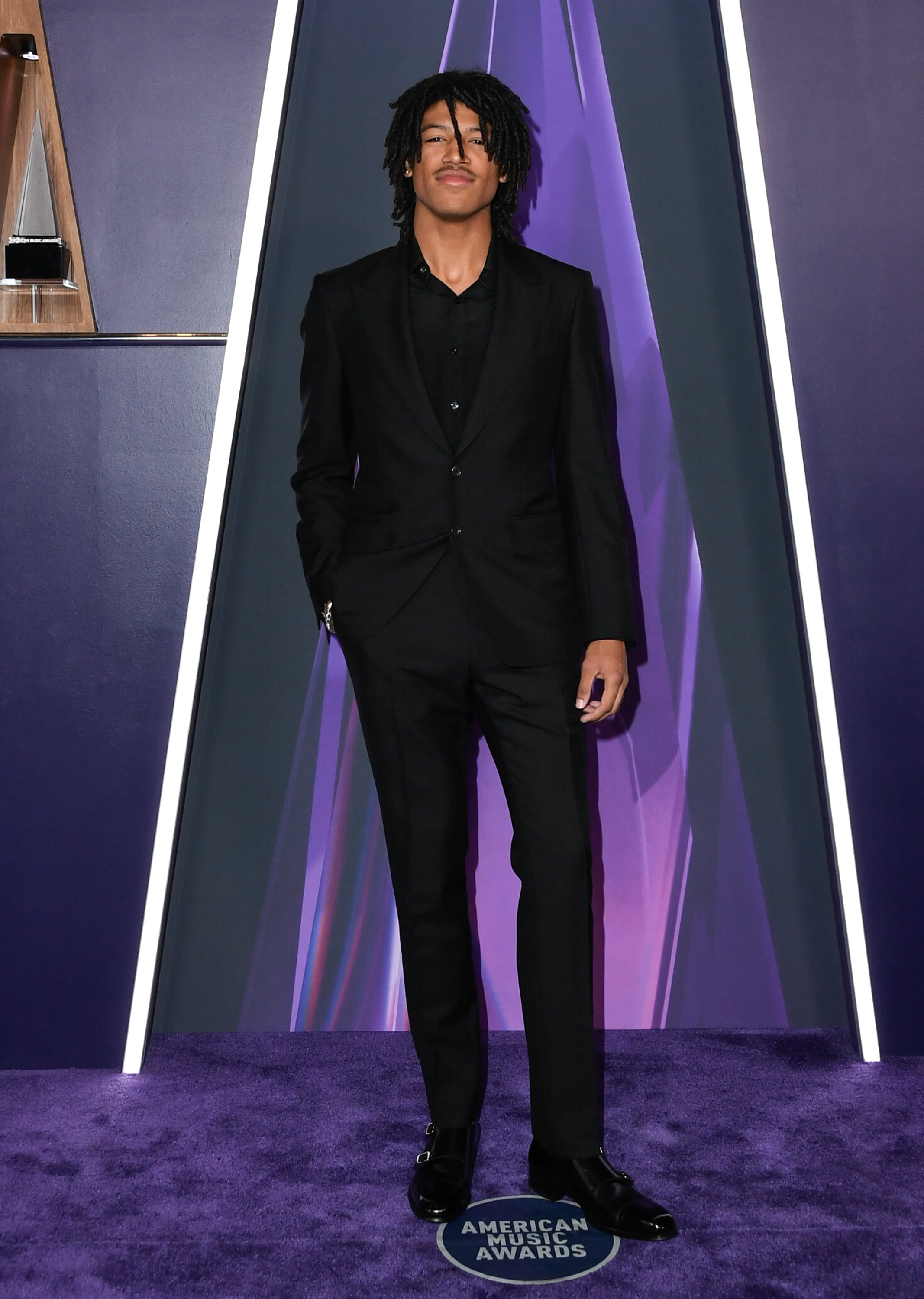 Henry Samuel stands with a relaxed stance and one hand in his pocket, meeting the camera with a composed expression as he poses solo on the purple carpet at the 2025 AMAs.