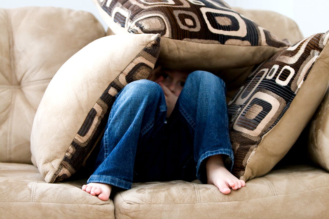 A picture of a young boy hiding his face under throw pillows. | Photo: Pexels.