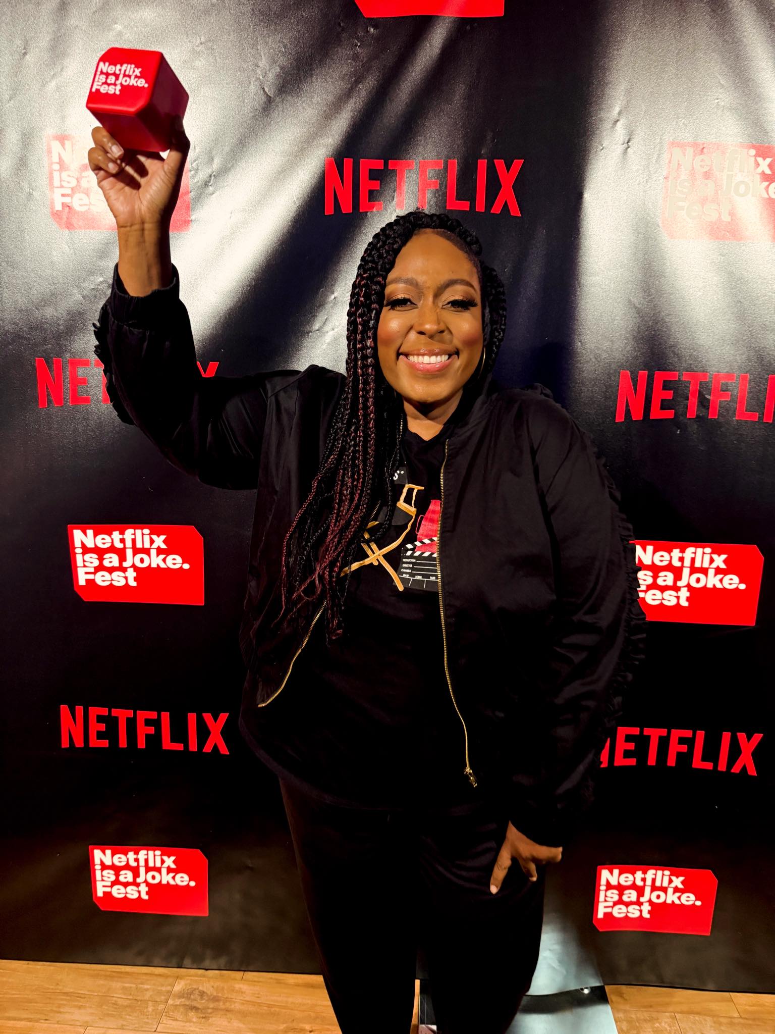 Loni Love appears at Netflix Is a Joke Fest, smiling widely as she raises a branded microphone against a black Netflix backdrop while dressed in a casual black jacket and pants. | Source: Facebook/Loni Love