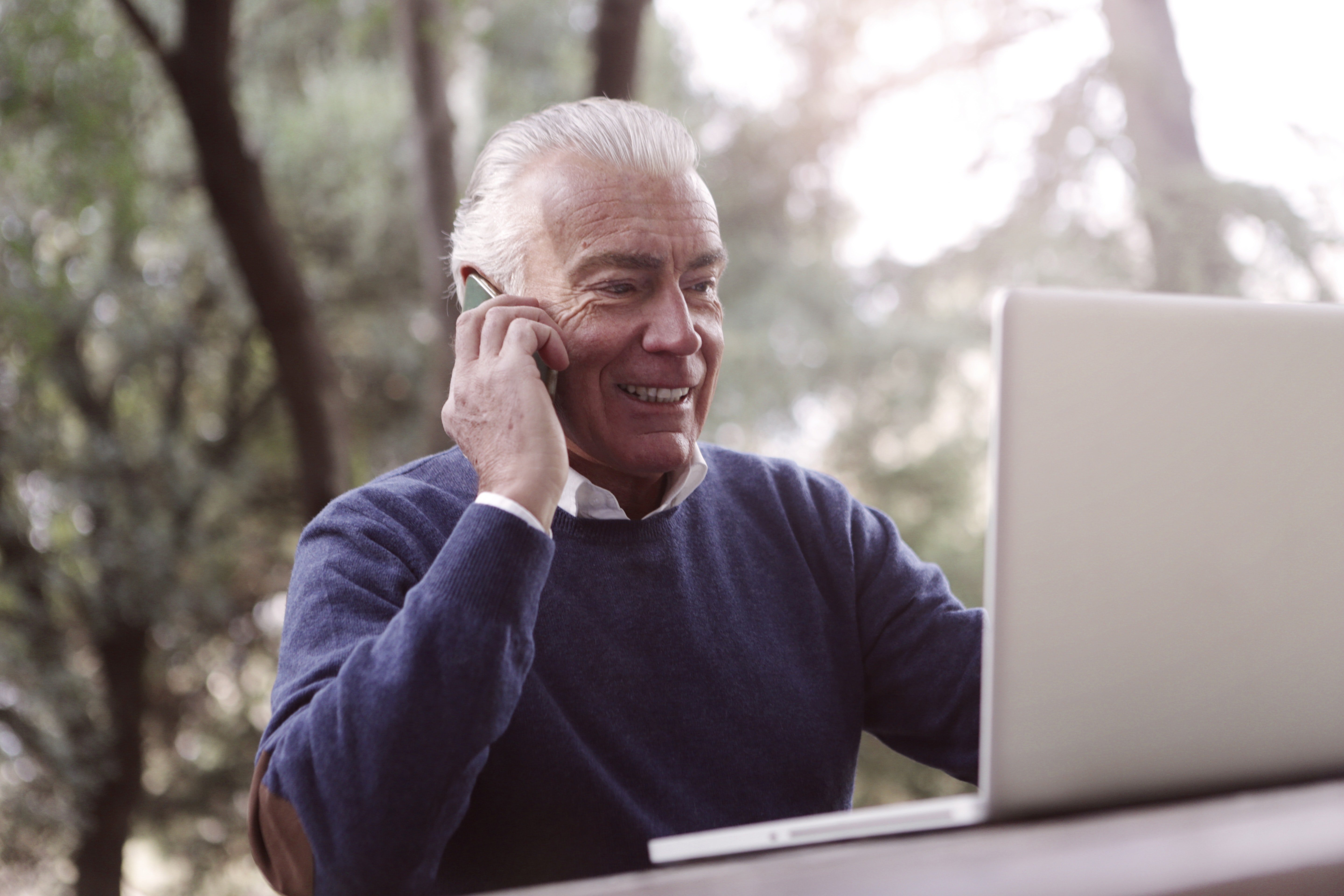 A smiling man on his cellphone with a laptop in front of him. | Photo: Pexels