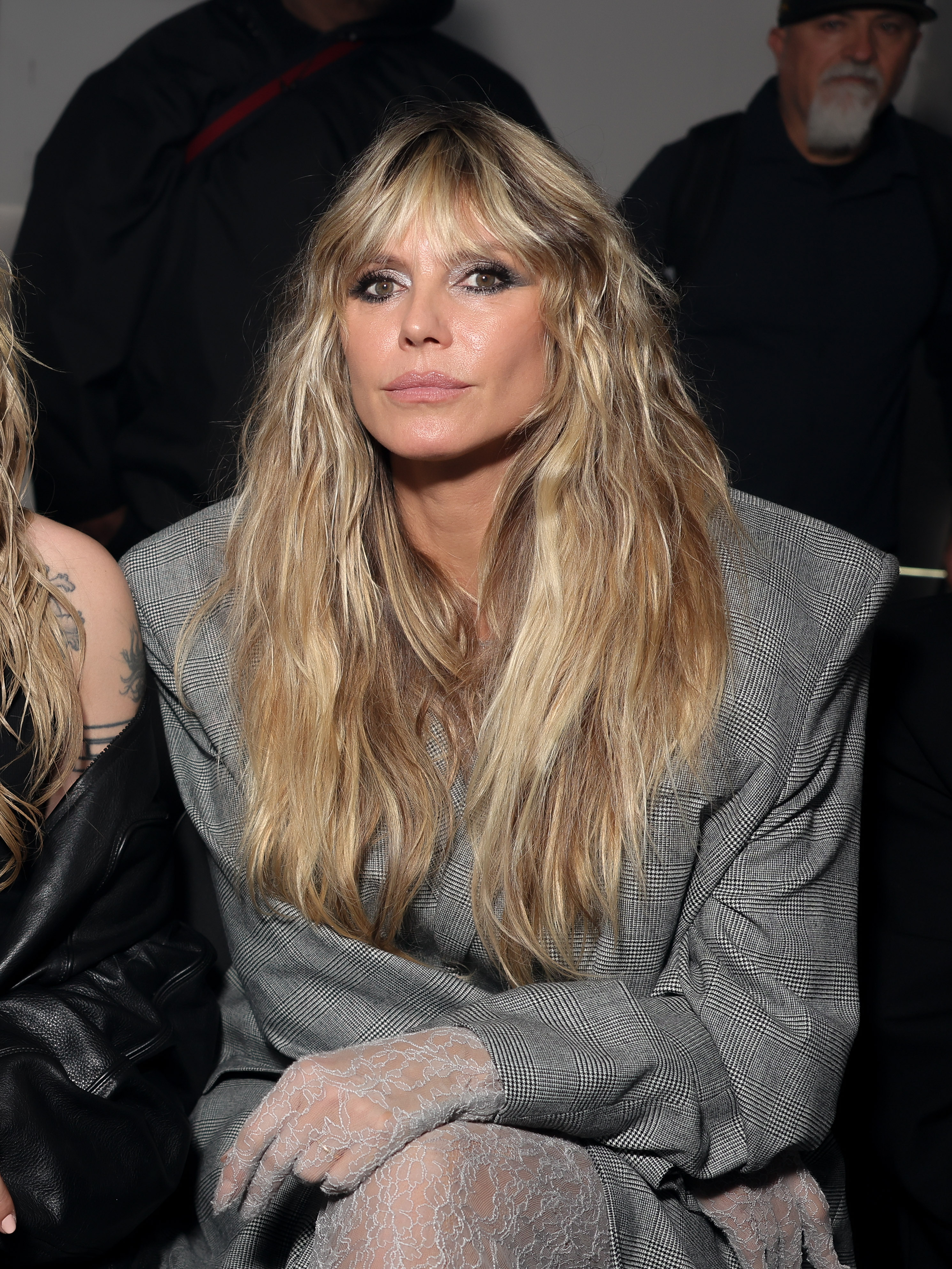 Heidi Klum sits watching the show in a gray blazer draped over her sheer outfit. Her blonde hair is full and cascading over her shoulders as she gazes ahead, looking as stunning as ever.