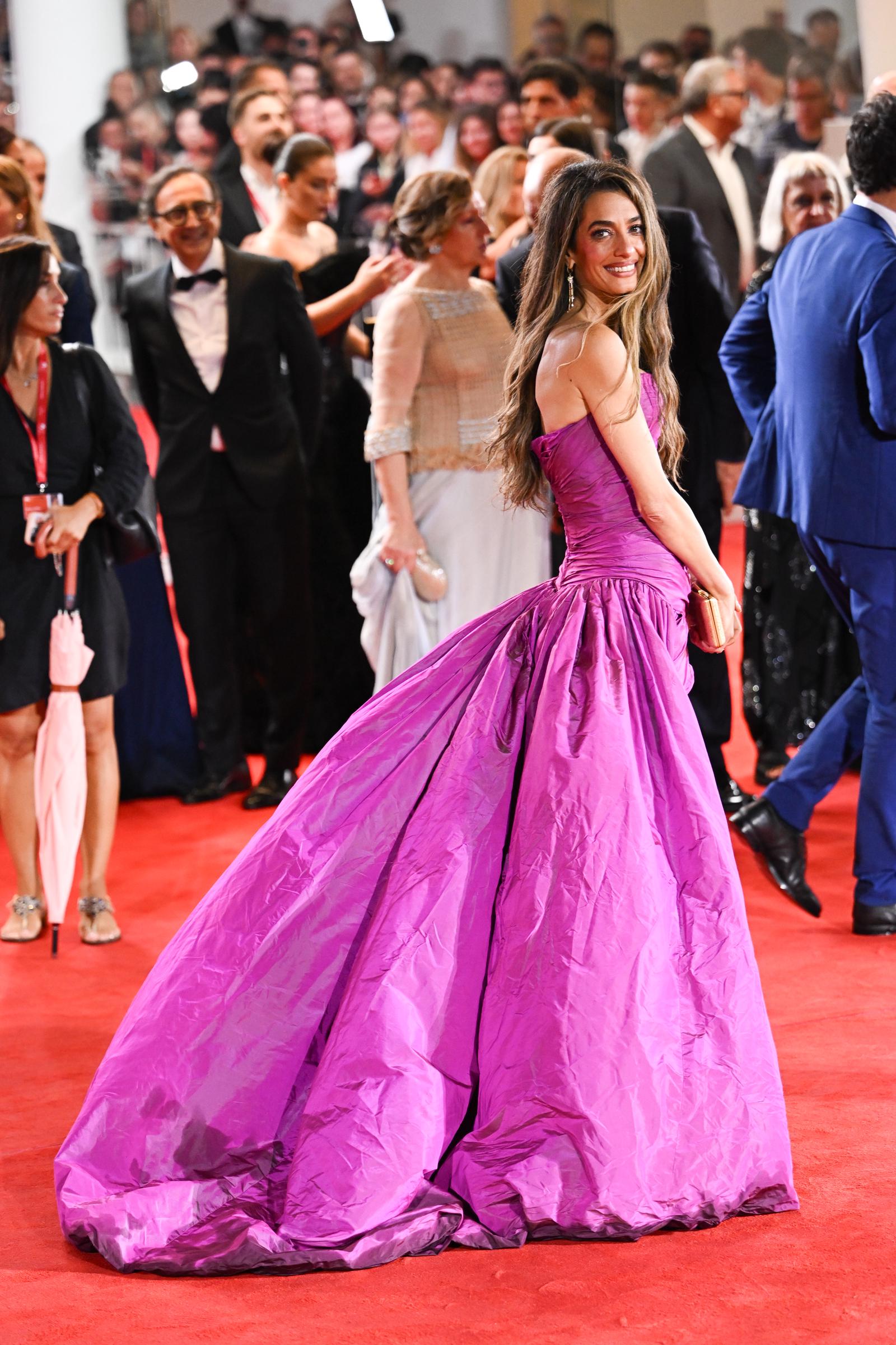 Amal Clooney glances over her shoulder with a bright smile, her voluminous fuchsia gown trailing behind her in dramatic folds. Surrounded by a crowd of guests and photographers, she holds her gold clutch as she makes her way through the red carpet.