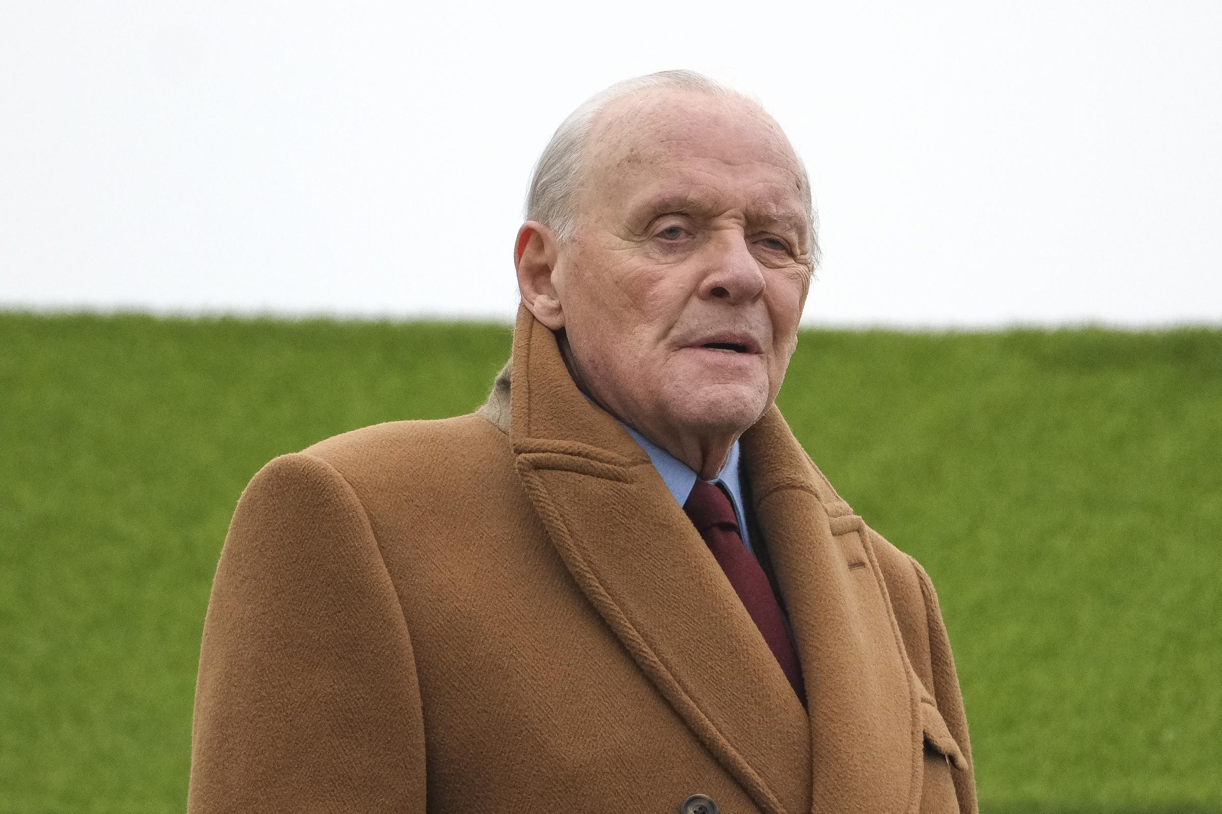 Anthony Hopkins is seen filming at Sandown Park Racecourse, wrapped in a rich camel overcoat that exudes quiet elegance. His poised expression and thoughtful gaze capture the essence of a man deep in character. Against the crisp green backdrop, he stands as a figure of grace and gravitas amid the English chill.