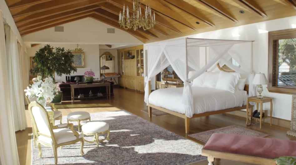 Exposed wooden ceiling beams and a striking brass chandelier crown this expansive primary suite, where a wooden canopy bed draped in airy white fabric stands as the central feature. Elegant gilded armchairs, a soft area rug, and a sitting area with a fireplace in the background give the high-ceilinged room a warm, luxurious atmosphere. | Source: YouTube/Architectural Digest