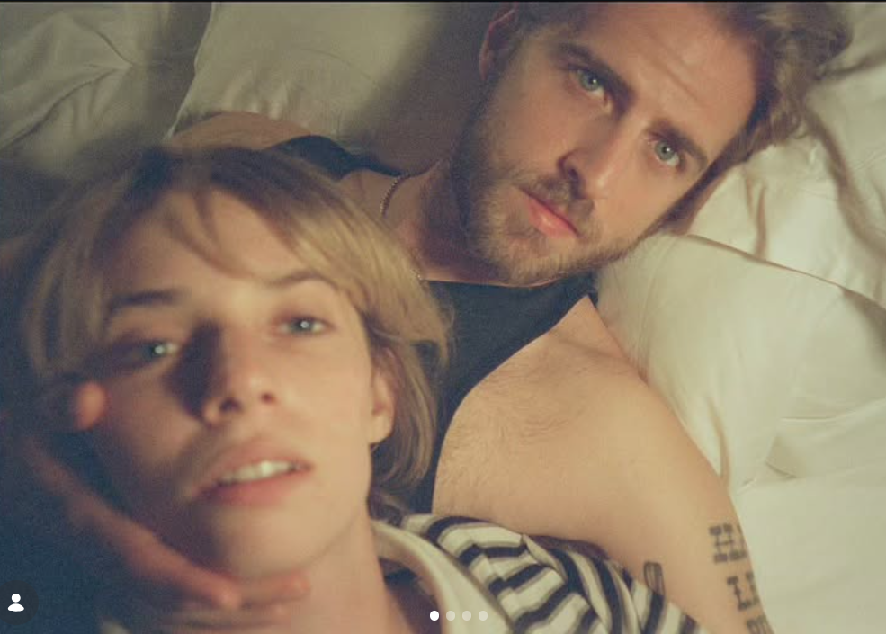 A softly lit, intimate close-up shows the couple lying in bed, Maya Hawke in the foreground and Christian Hutson gazing at the camera behind her. | Source: Instagram/maya_hawke