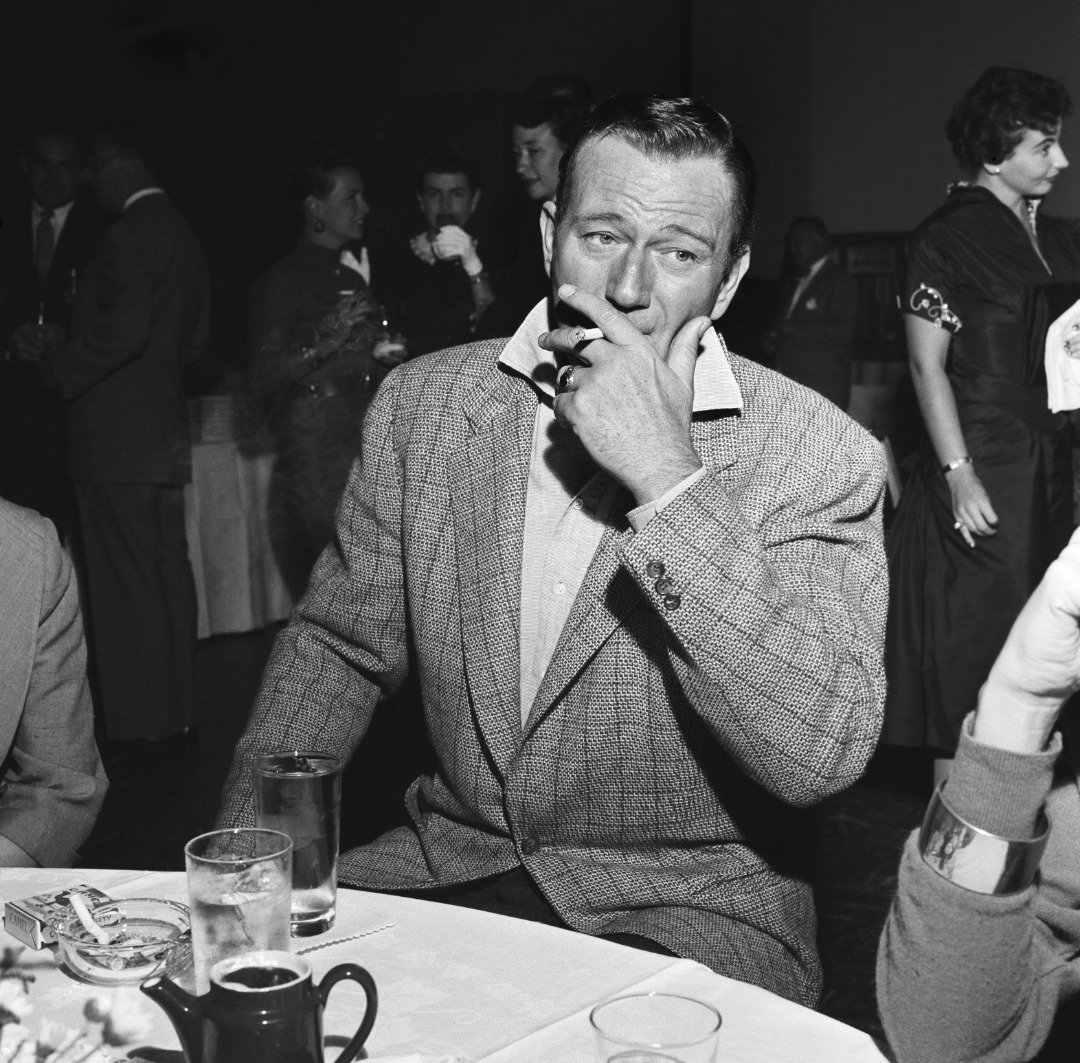 John Wayne smokes a cigarette at a party for Milton Berle on September 26 1955 in Los Angeles, California. | Source: Getty Images