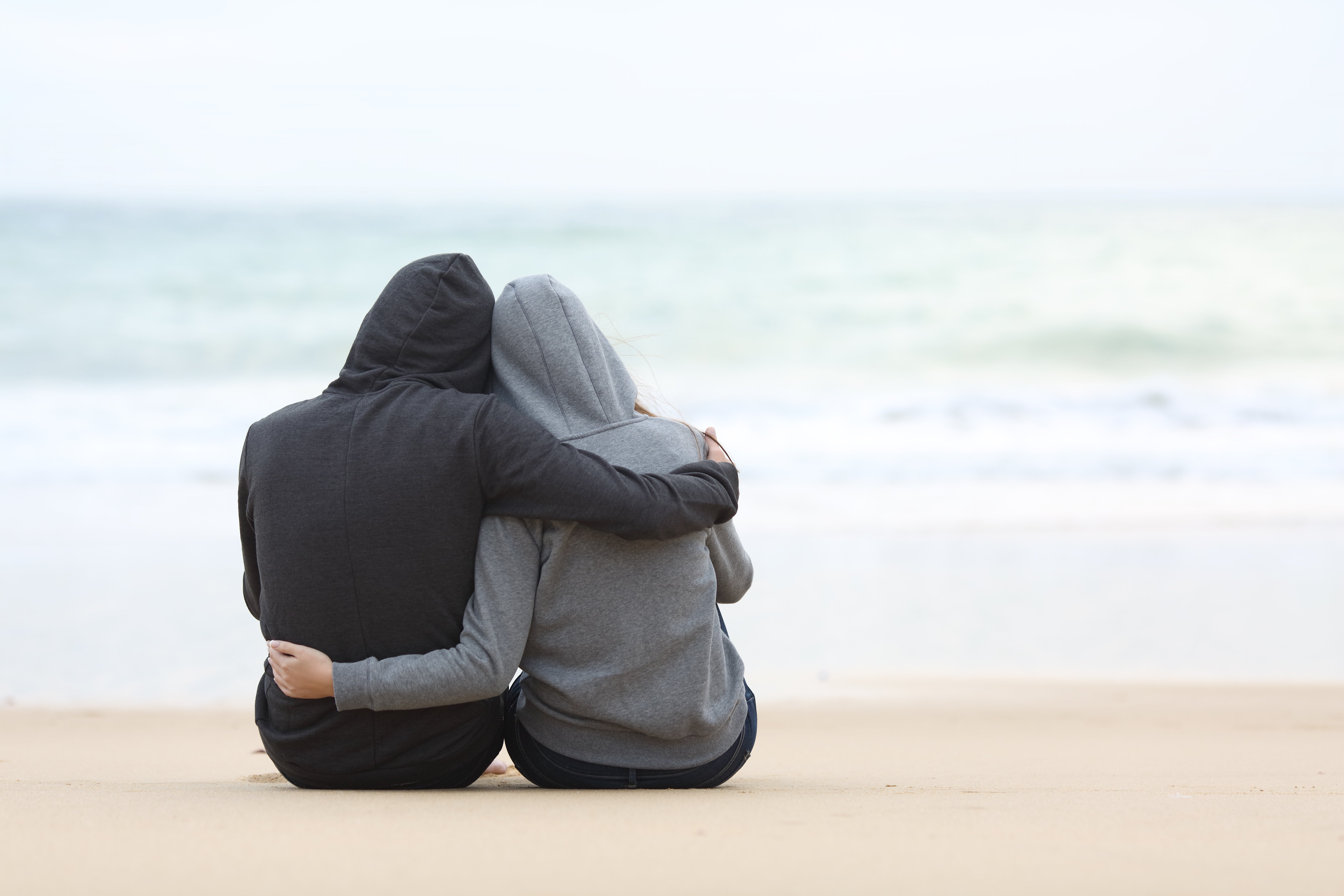 A rear view of two people hugging and watching the sea while sitting on the beach sand on a rainy day | Source: Shutterstock