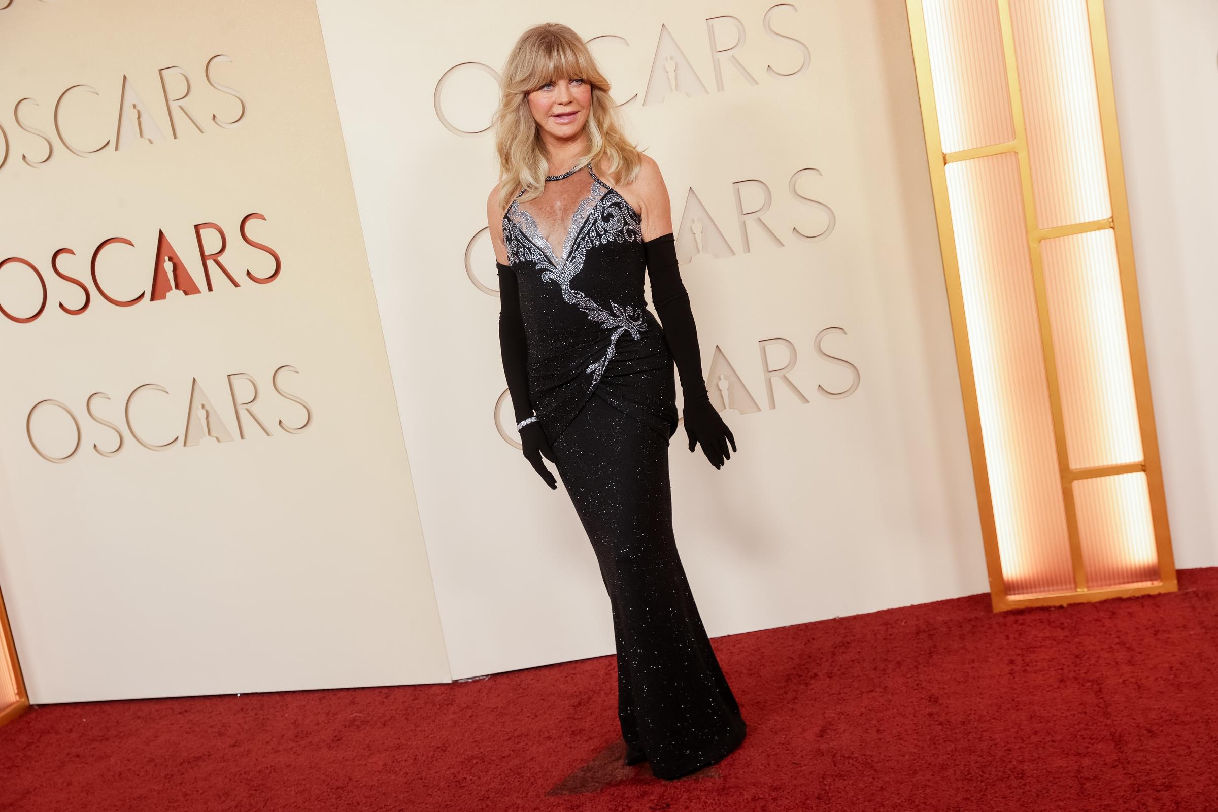 Goldie Hawn steps onto the Oscars red carpet in a sleek black gown with shimmering silver detailing and long opera gloves, showcasing the timeless elegance that has defined her Hollywood career.