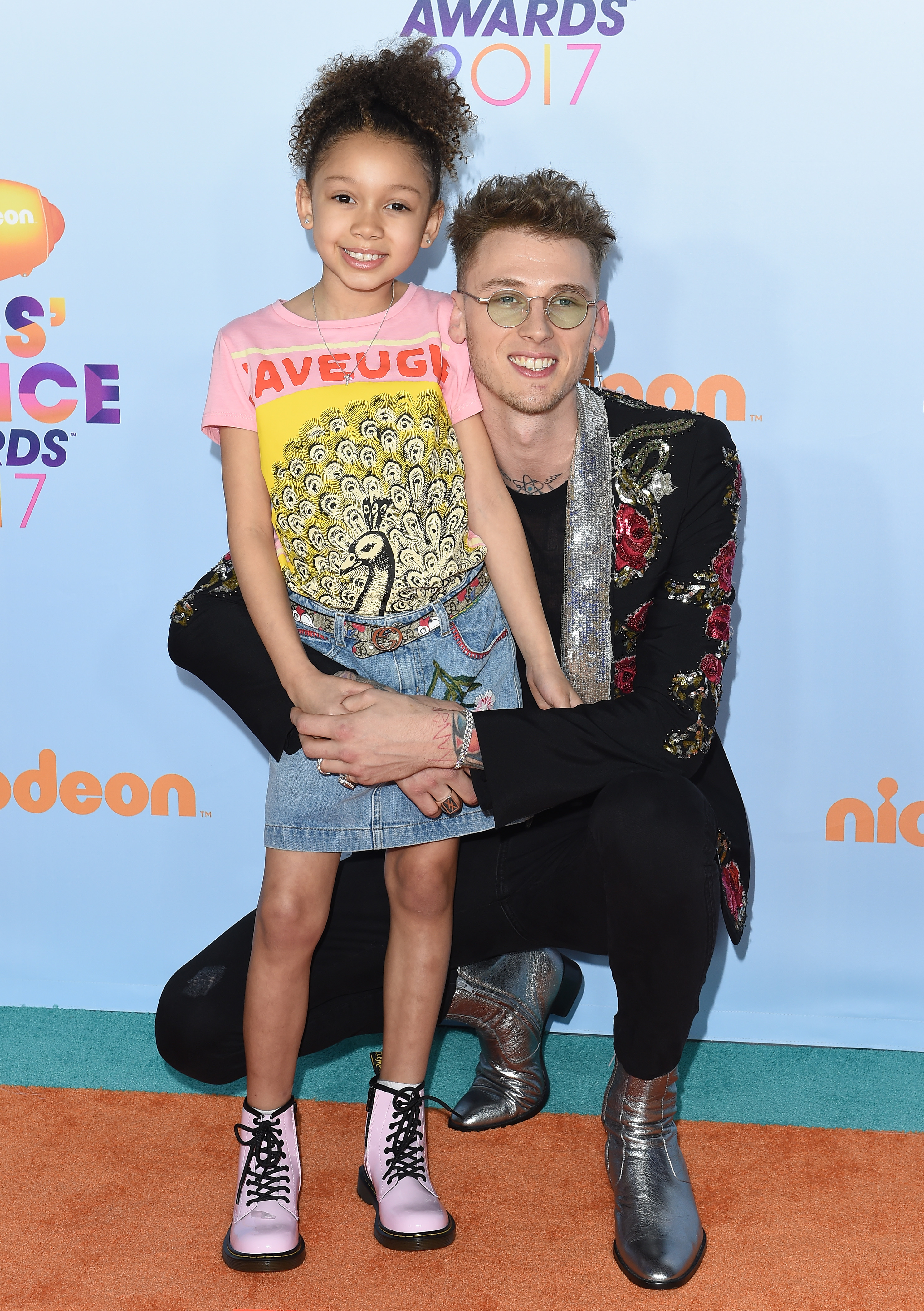 Machine Gun Kelly kneels beside Casie on the 2017 Kids' Choice Awards carpet, wrapping both arms around her waist as she stands smiling. The pair pose cheerfully for the cameras.
