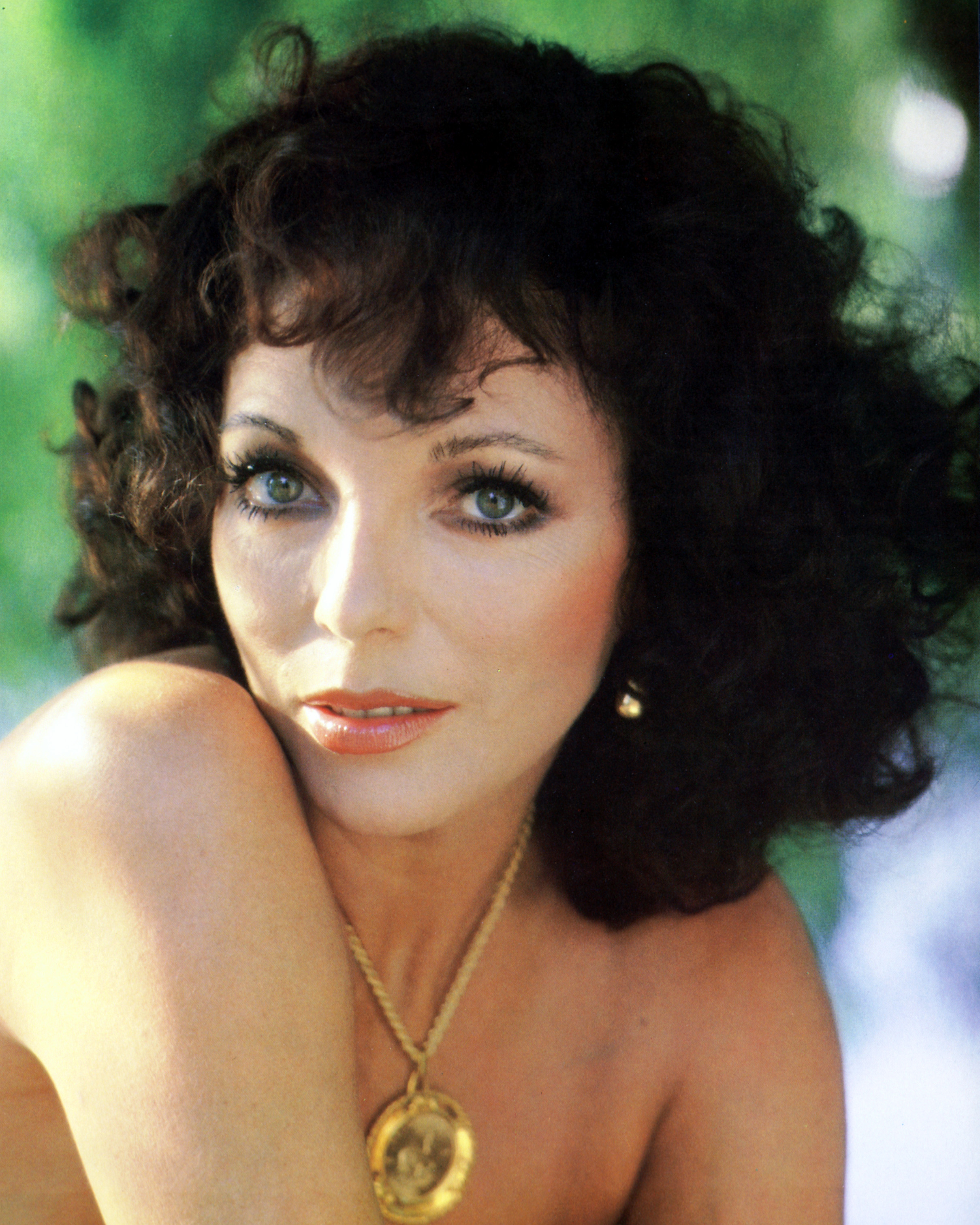 Bathed in soft natural light and framed by a lush green backdrop, Joan Collins embodied effortless 1970s elegance in this portrait taken circa 1970. Her voluminous curls, dewy complexion, and lightly glossed lips lent a fresh, sun-kissed appeal, while a bold gold medallion necklace and matching earrings hinted at the era's growing taste for statement glamour.