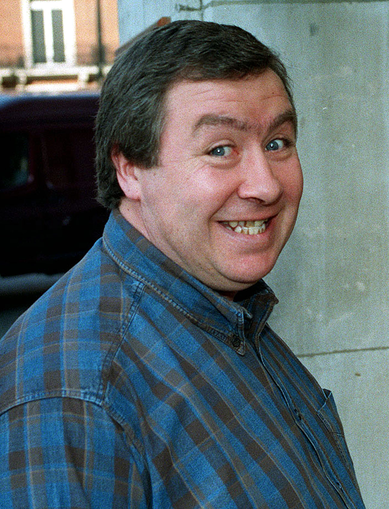 Actor Gregor Fisher smiling toward the camera in a blue plaid shirt, captured in a candid moment early in his career.