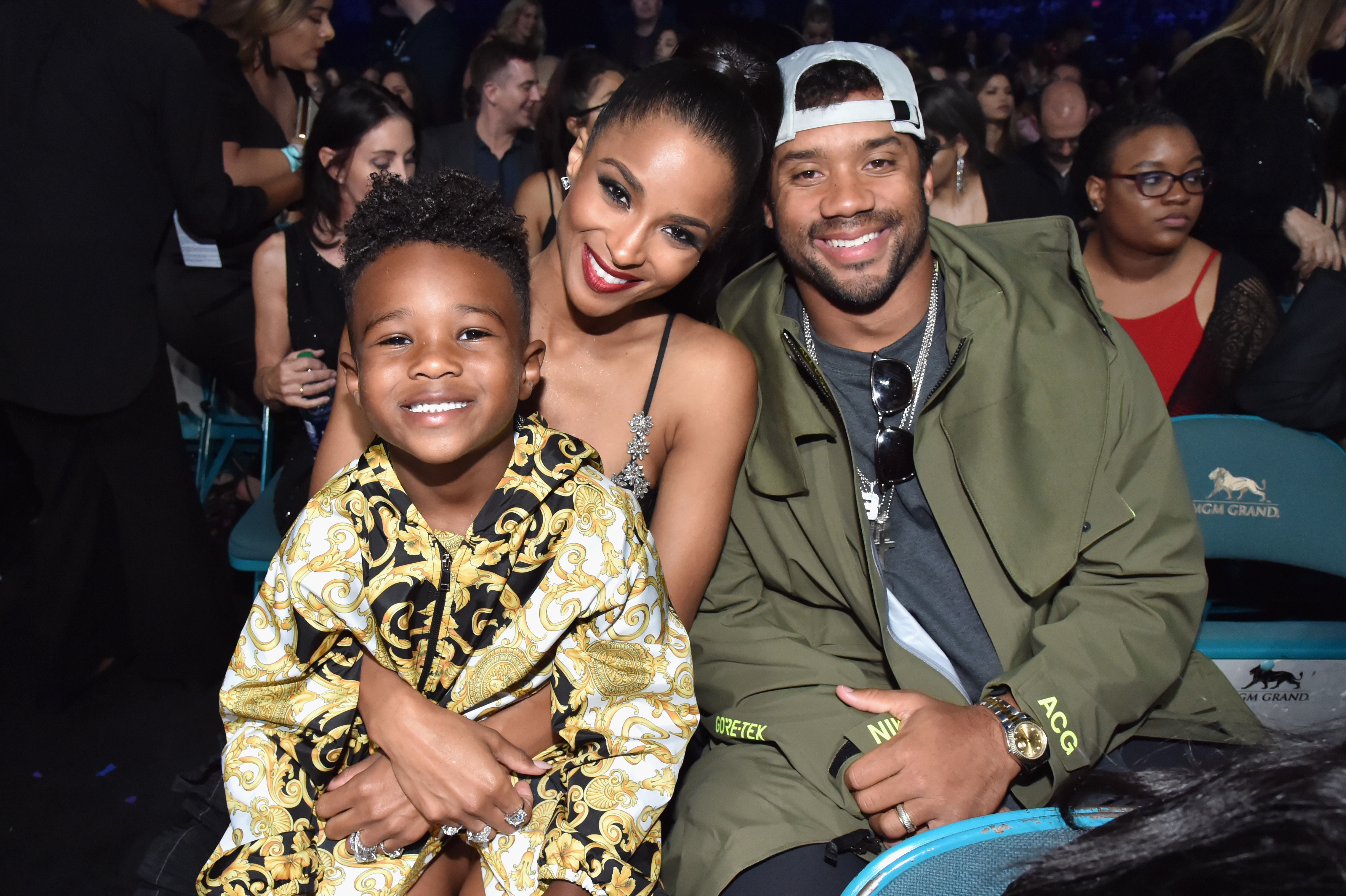 Seated together, Ciara leans in with a bright, joyful smile while Russell Wilson sits beside her with a relaxed, friendly grin. A young Future Zahir Wilburn sits in front of them, smiling toward the camera, adding a warm, intimate family touch to the scene.
