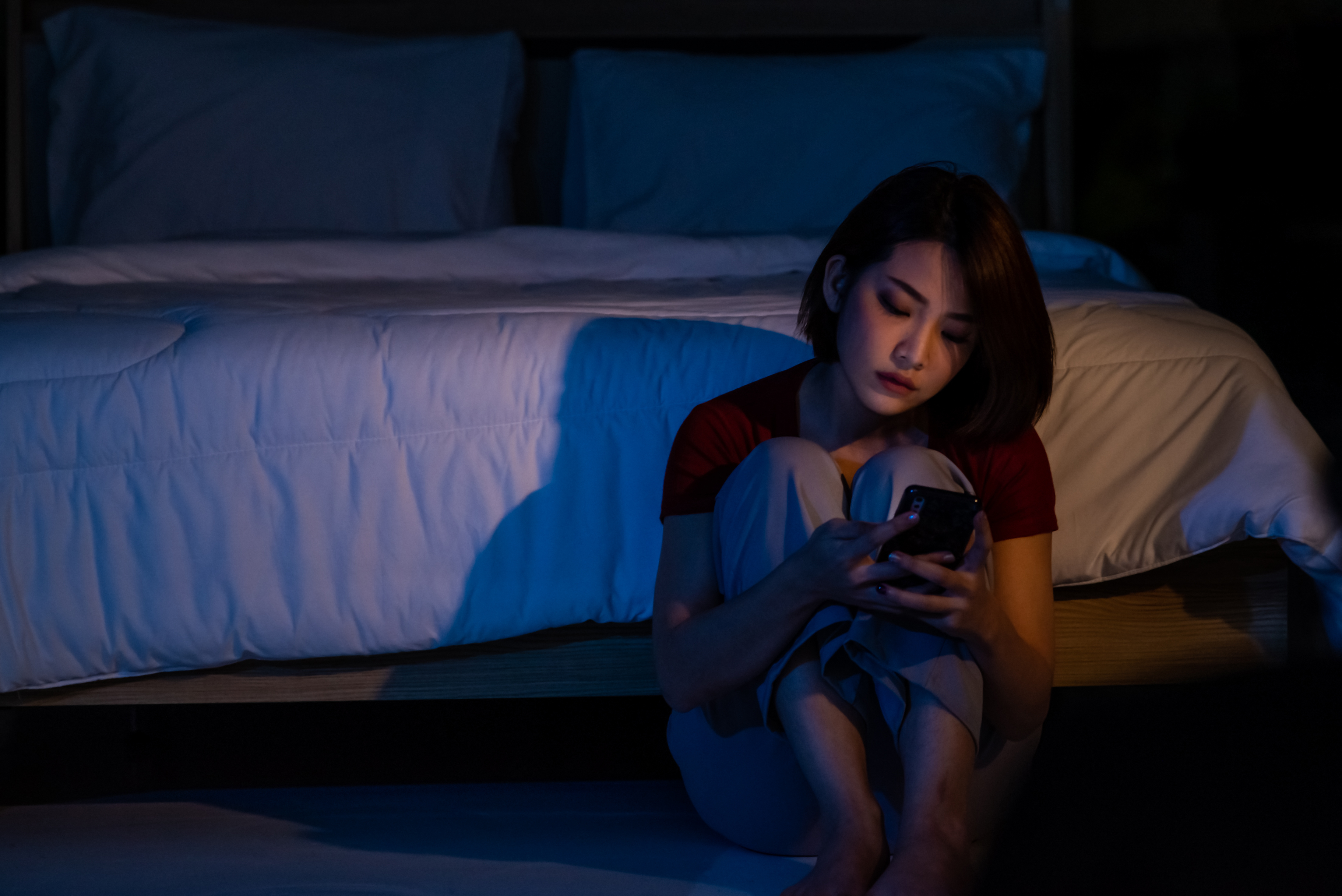 Depressed young beautiful Asian woman sitting alone on bedroom floor with looking at smartphone. Stressed insomnia sad woman having life problems. Mental health, healthcare and social issue concept | Source: Getty Images