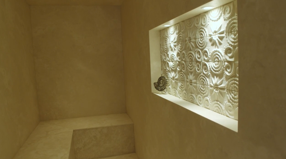 Travertine stone seating and smooth warm-toned walls define this private steam room, highlighted by a glowing illuminated niche showcasing intricate carved tilework and a decorative nautilus shell. | Source: YouTube/Architectural Digest