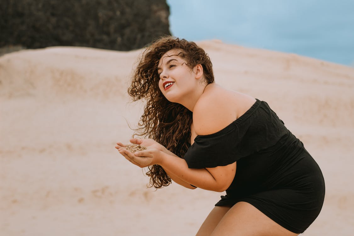 Mujer posando de rodillas sobre la arena mientras usa un vestido de playa corto de color negro. | Foto: Pexels