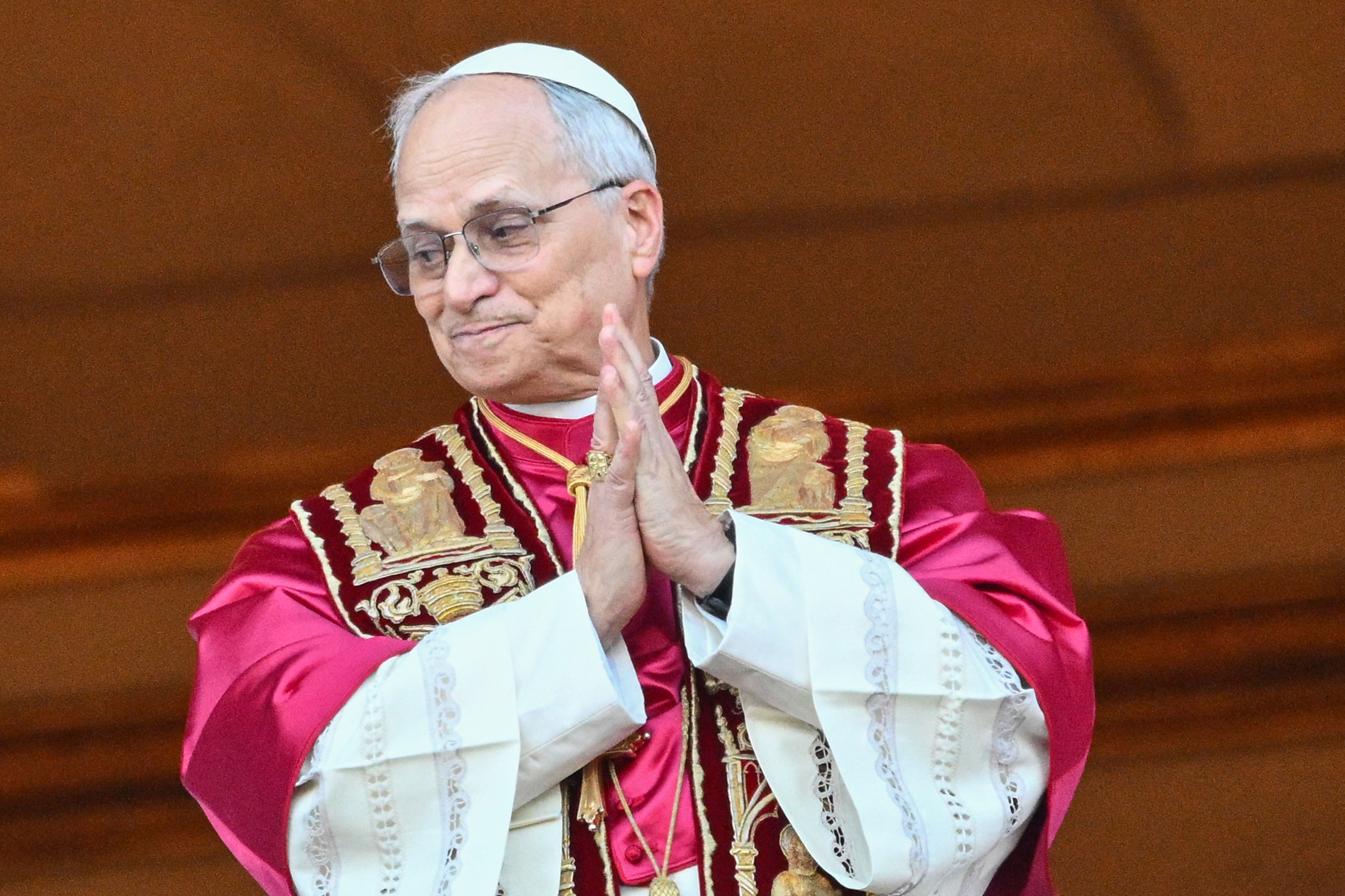 Pope Leo XIV presses his hands together with a soft smile, his crimson and gold vestments gleaming under the lights of the basilica balcony