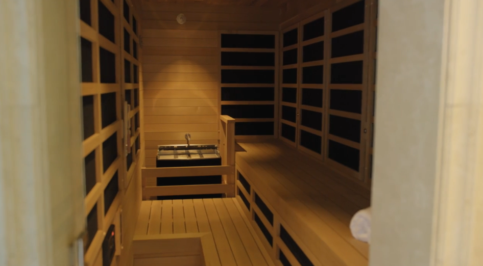 Natural wooden slatted panels and bench seating line this custom built-in sauna, centered around a traditional stone heating stove. Integrated warm lighting soft-lights the timber walls, creating a relaxing Scandinavian-style wellness space inside the residence. | Source: YouTube/Architectural Digest