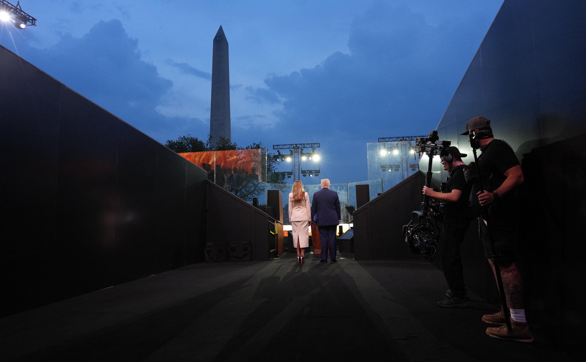 Donald and Melania Trump walk down a backlit corridor toward the stage, the Washington Monument glowing in the distance. Camera crews capture their approach during the U.S. Army’s 250th anniversary parade on June 14, 2025.