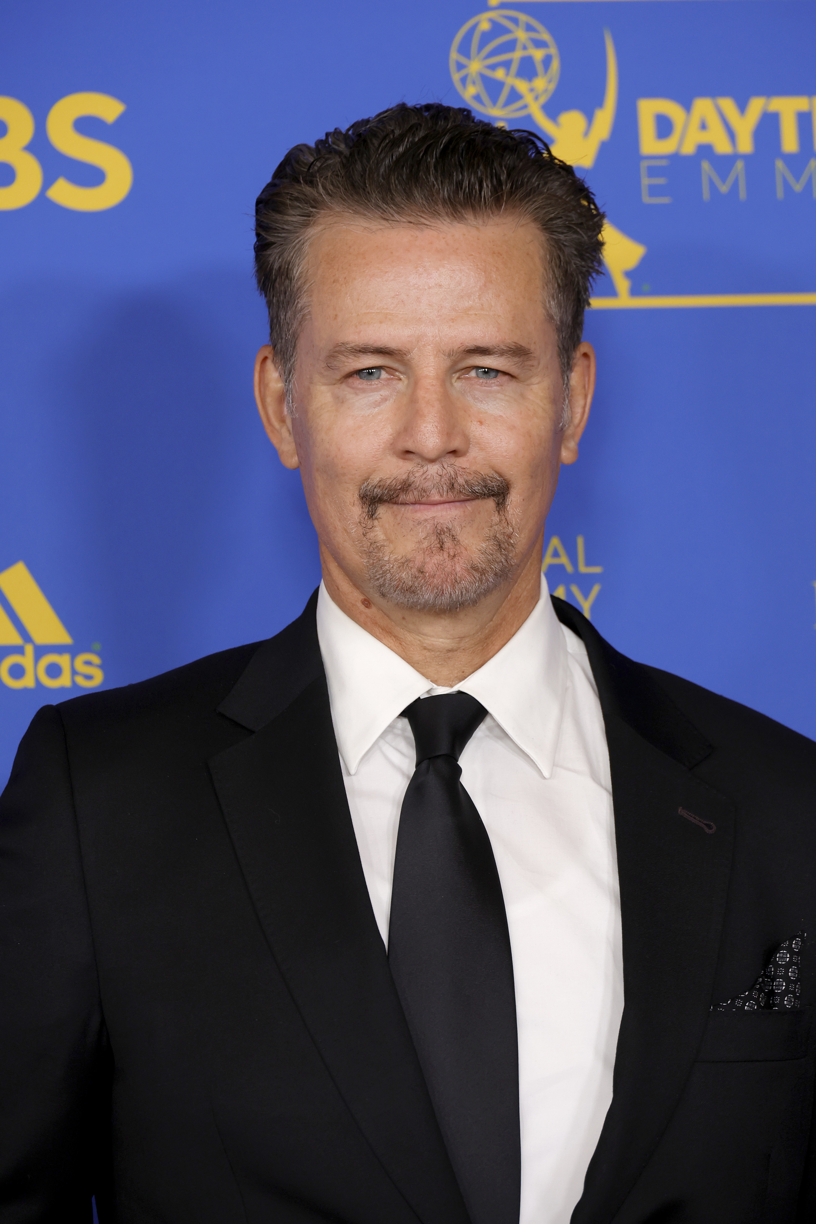 Ted King appears at the Daytime Emmy Awards in 2022, dressed in a classic black suit and tie with a white shirt. His neatly styled hair and trimmed goatee complement the formal setting, while the Emmy backdrop reinforces the celebratory nature of the occasion.