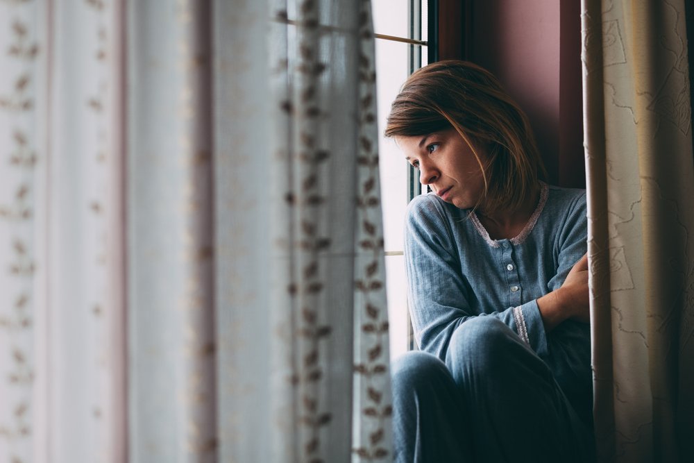 Une jeune femme déprimée et culpabilisée, assise à la fenêtre, regardant dehors. | Shutterstock