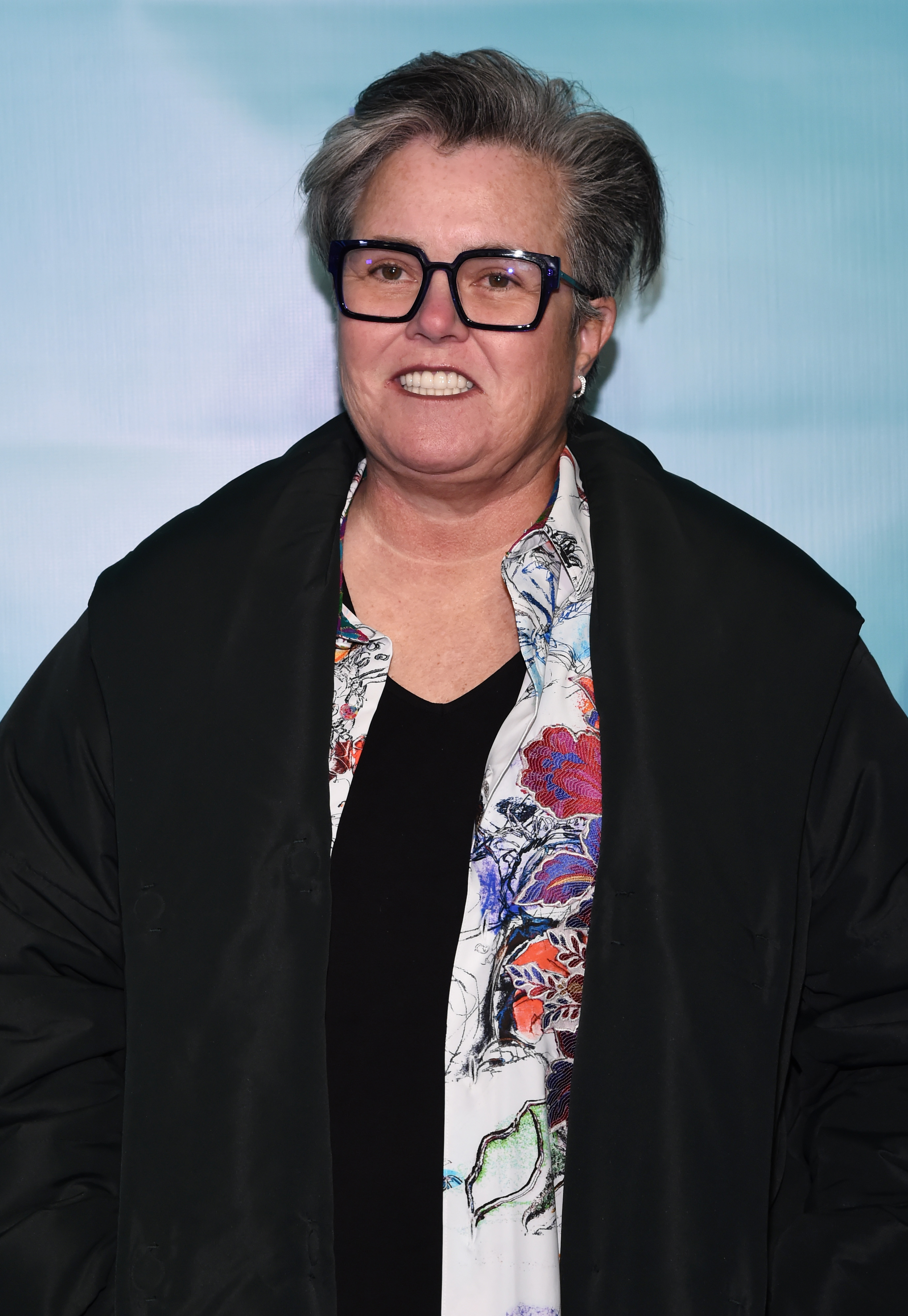 At the "Jagged Little Pill" Broadway premiere on December 5, 2019, Rosie O'Donnell wears a colorful blouse and dark jacket, flashing a cheerful smile with brushed-back hair and her signature dark frames.