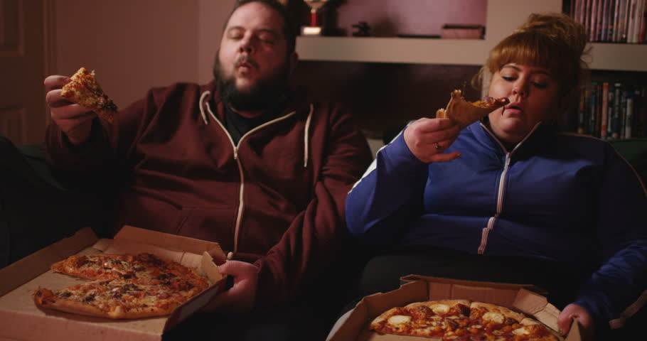 An overweight couple, eating pizza in their living room | Source: Shutterstock.com