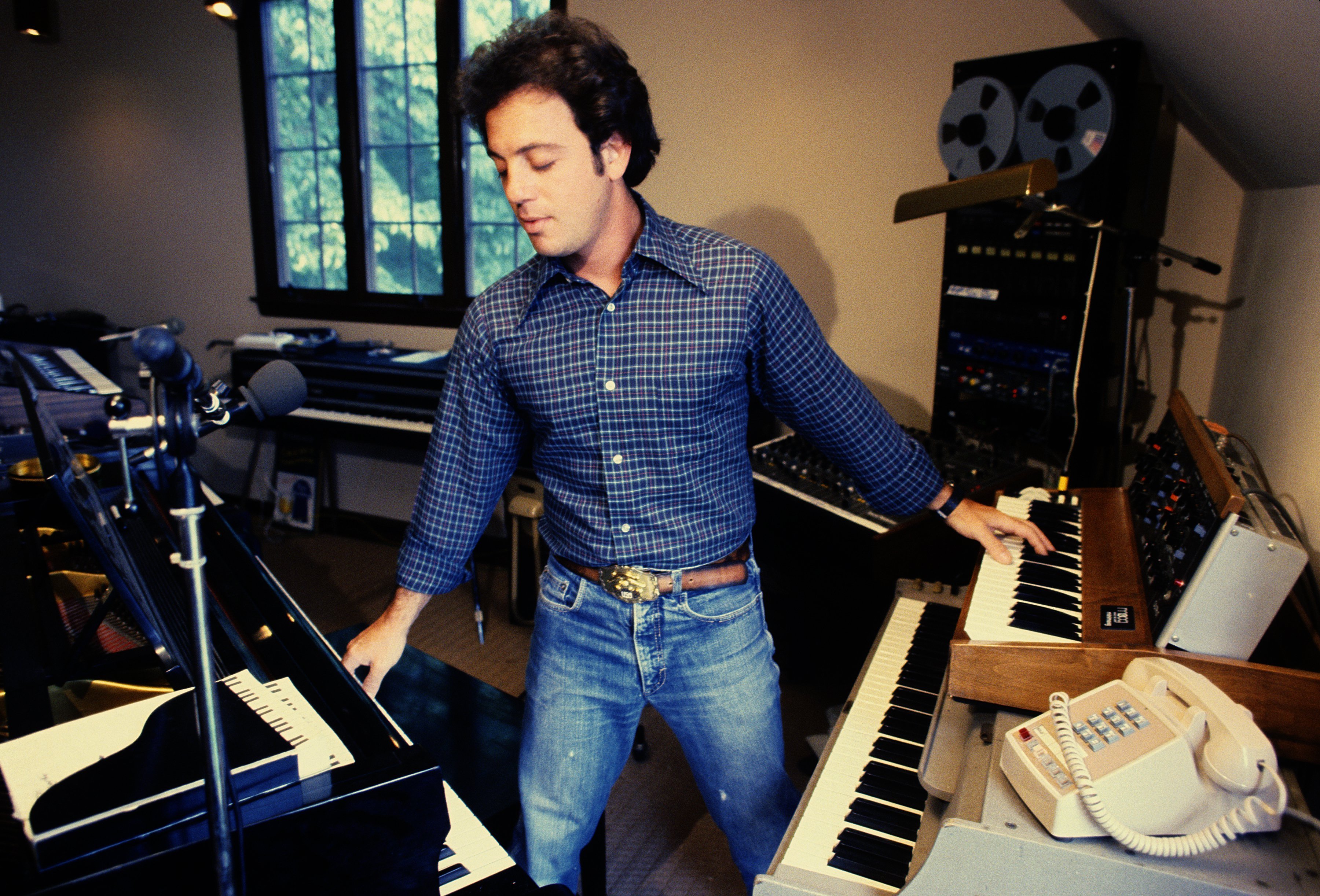 Composer Billy Joel wearing a checkered shirt paired with jeans while performing in his home studio at his Los Angeles home in 1984. / Source: Getty Images