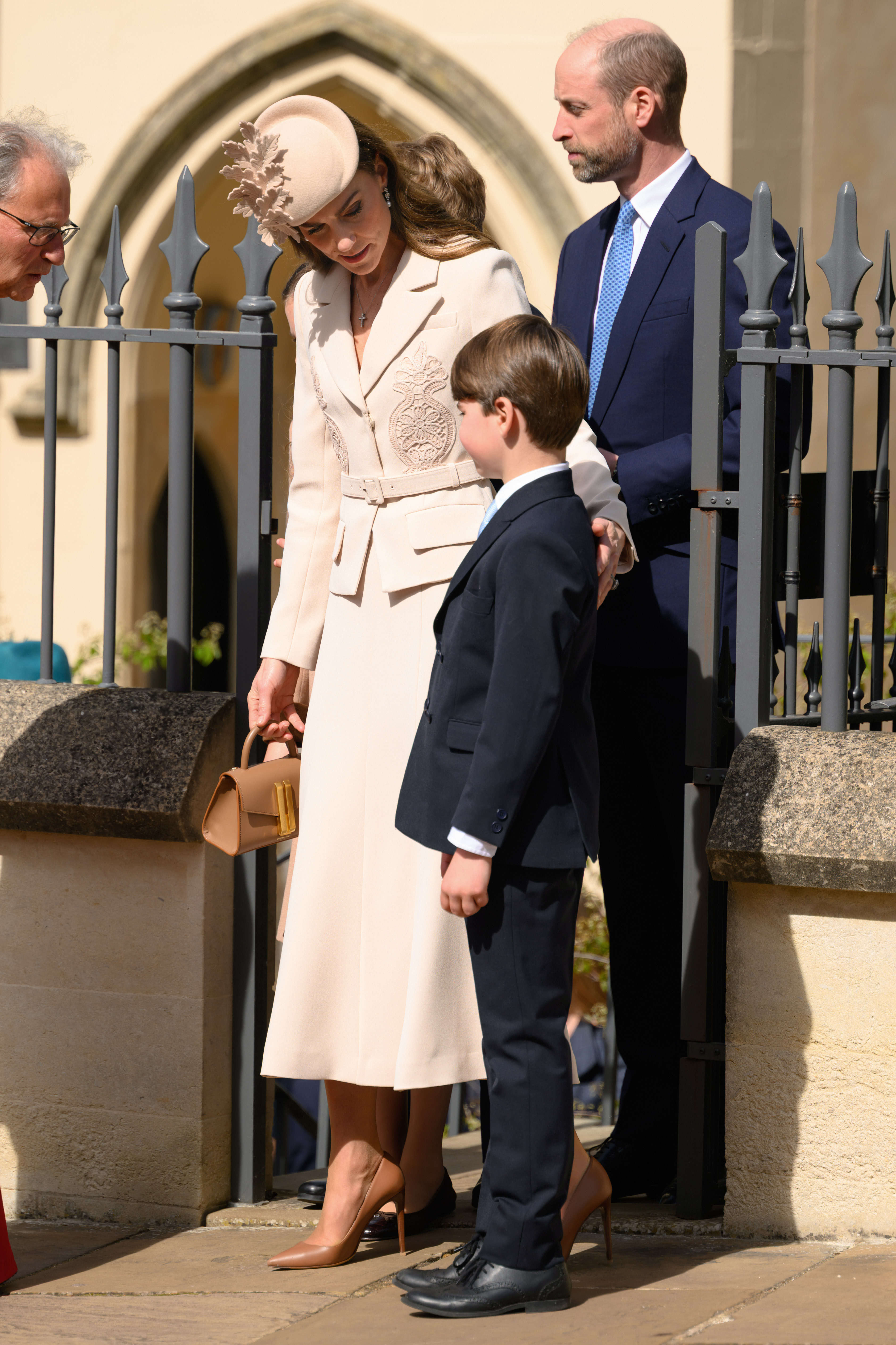 Outside St George's Chapel, the Princess of Wales leans down toward Prince Louis, 7, in a quiet moment between the two, while the Prince of Wales stands just behind them in his navy suit and pale blue tie. The full length of the Princess of Wales's cream ensemble is visible here, from the structured blazer and calf-length skirt to her Ralph Lauren pumps and toffee-coloured DeMellier bag, held loosely at her side.