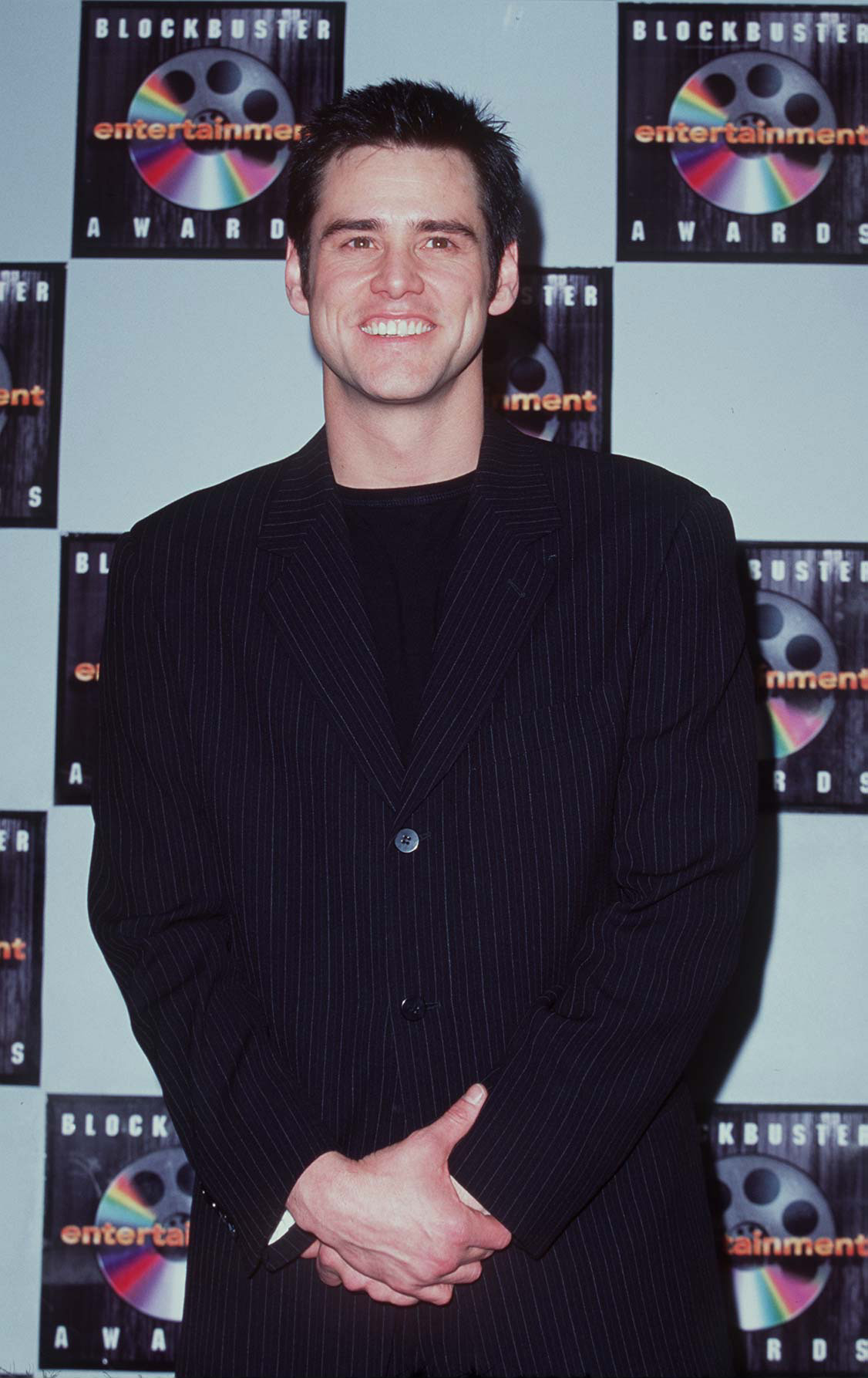 Smiling broadly beneath spiked hair, Jim Carrey poses in a sharp, dark pinstripe suit layered over a simple black tee at The 2nd Annual Blockbuster Awards. With his hands loosely clasped and posture relaxed, he radiates the easygoing confidence of a comedy heavyweight in his prime.