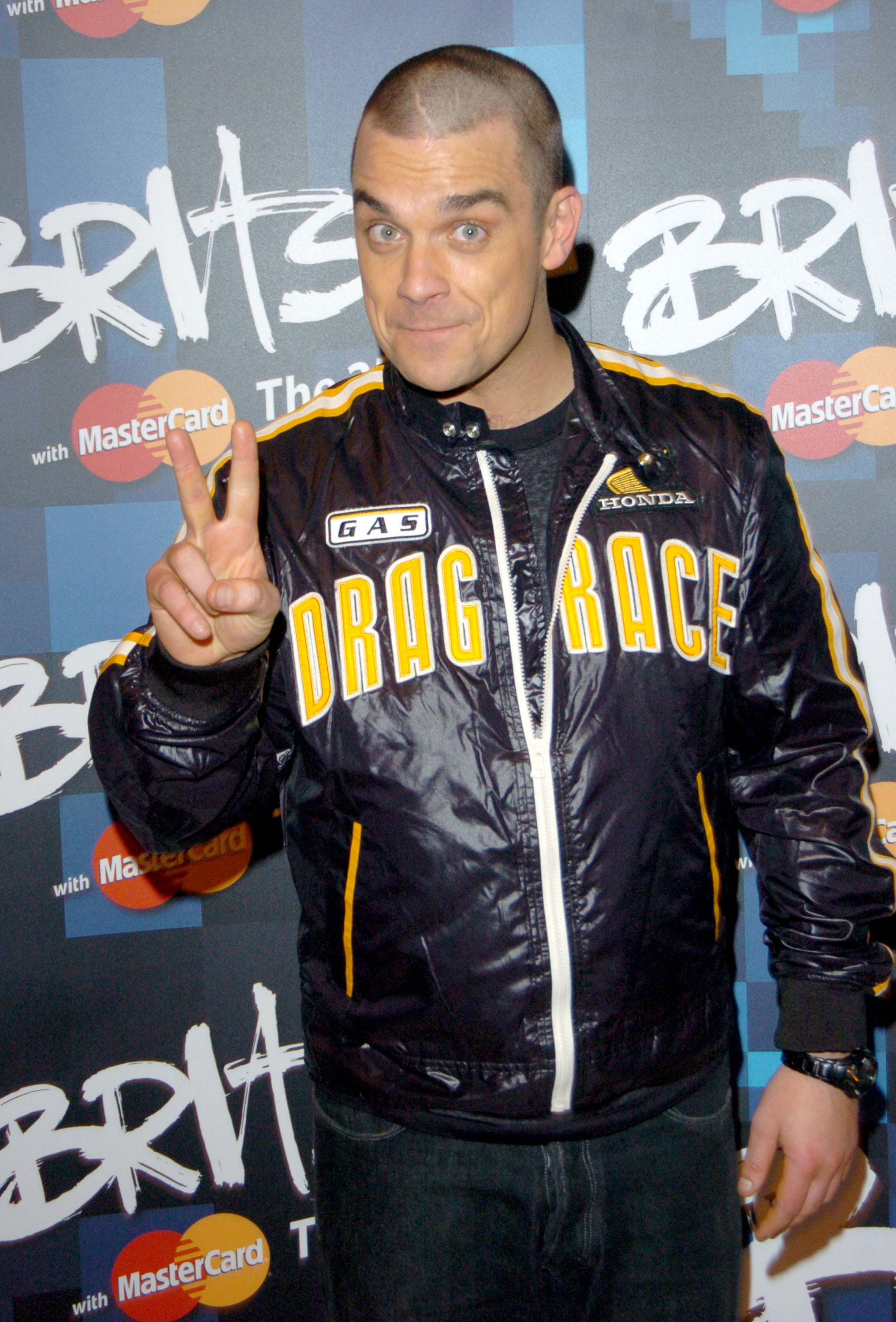 Robbie Williams posed at the 2005 Brit Awards flashing a peace sign in a bold "Drag Race" jacket. With a freshly shaved head and playful energy, he once again proved his knack for mixing mischief with style on the red carpet.