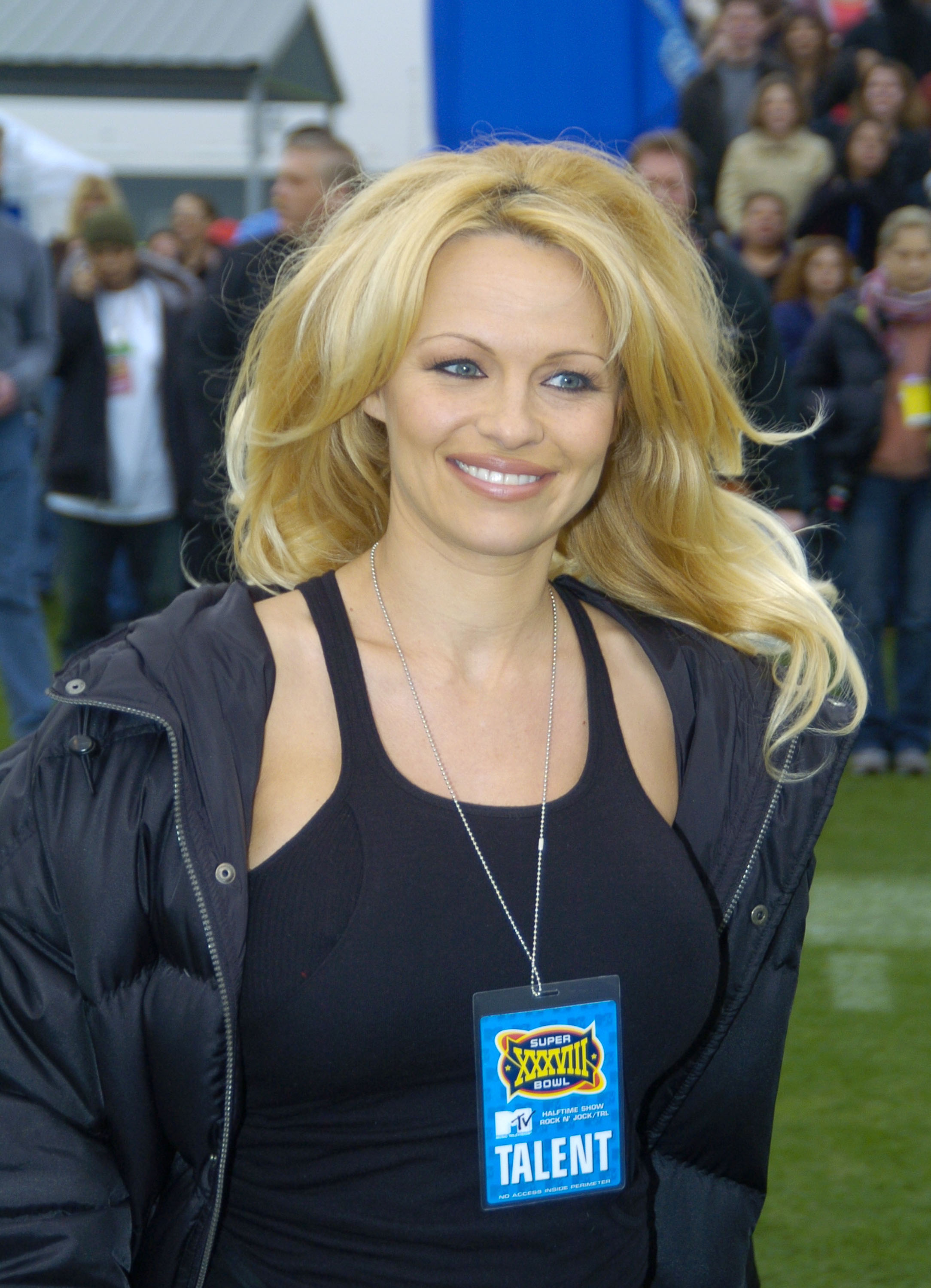 Pamela Anderson, with voluminous, wavy blonde hair, wears a black tank top and a black puffer jacket over a silver necklace with a "Super Bowl XXXVIII" talent pass attached, during MTV's "TRL at the Super Bowl" on January 31, 2004, at Reliant Stadium in Houston, Texas.