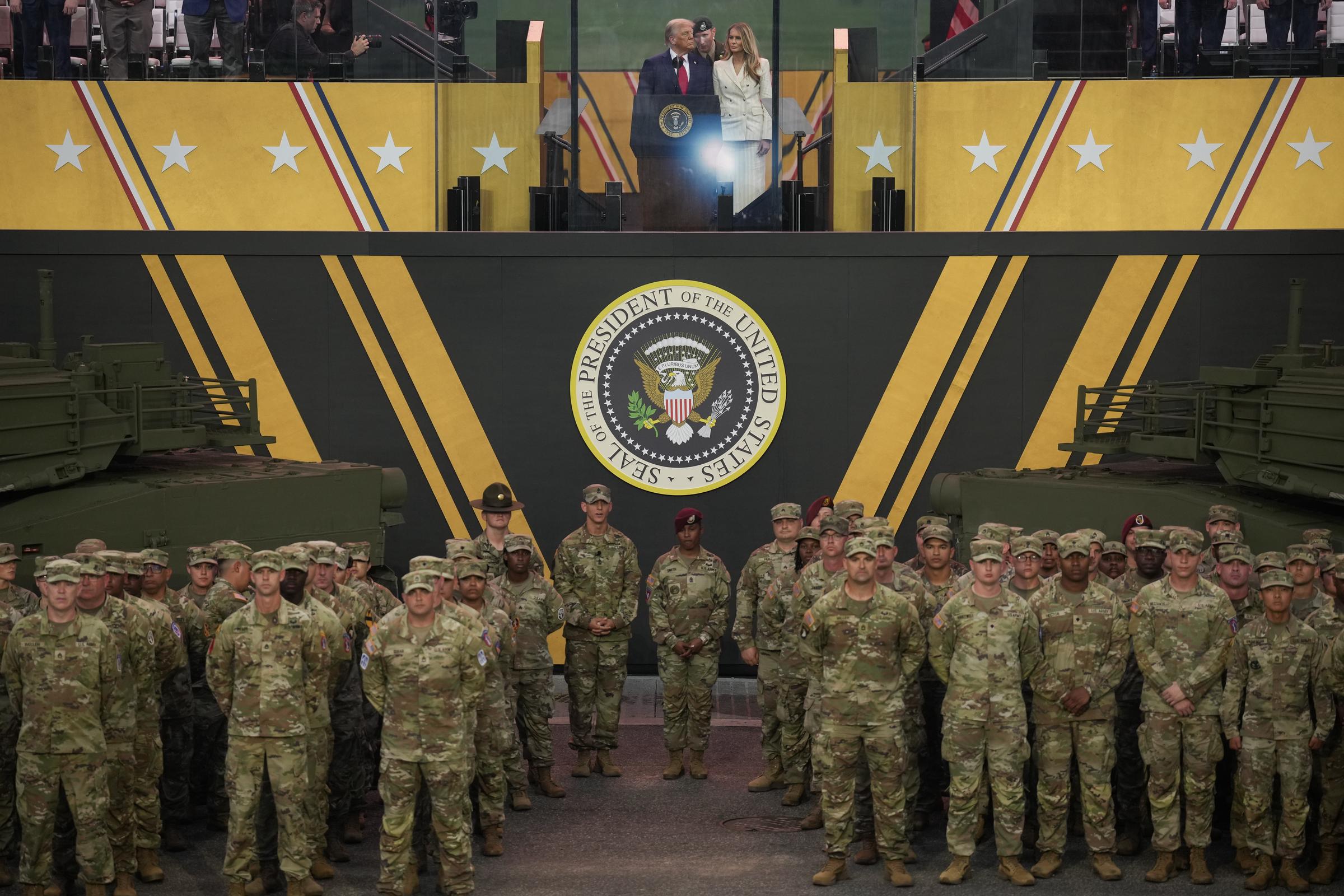 Donald and Melania Trump stand on stage above rows of U.S. soldiers at the conclusion of the parade. Framed by tanks and the presidential seal, the image captures the couple during a formal farewell moment before the assembled troops.