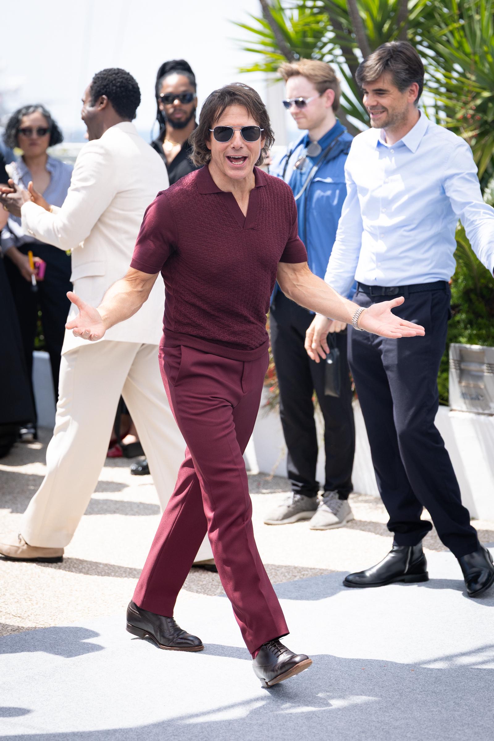 Tom Cruise steps into the spotlight with a lively gesture during the "Mission: Impossible – The Final Reckoning" photocall. Dressed in a burgundy ensemble and dark sunglasses, he engages with photographers and fans in classic Cruise fashion.