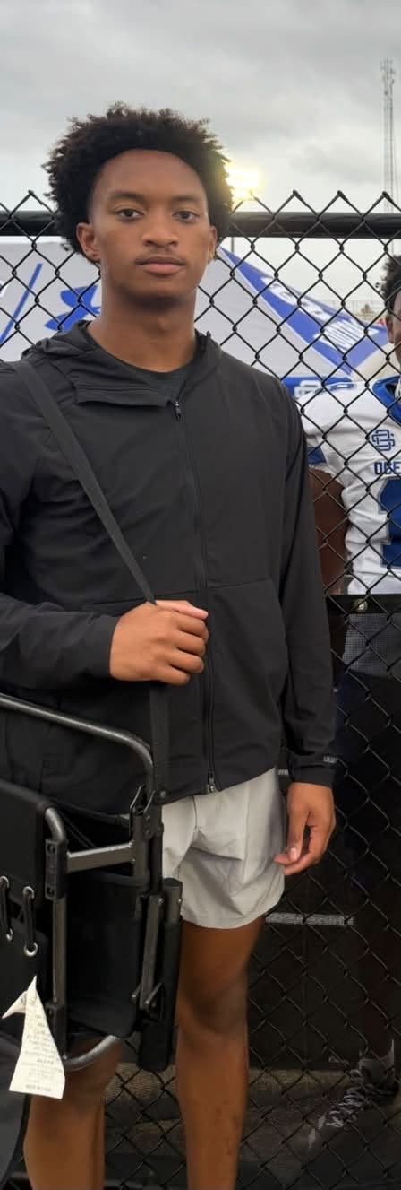 Dressed down in a black jacket and shorts, Nolan stands near the fence line at a game, camera bag slung over one shoulder. | Source: Facebook/christine.wonsley