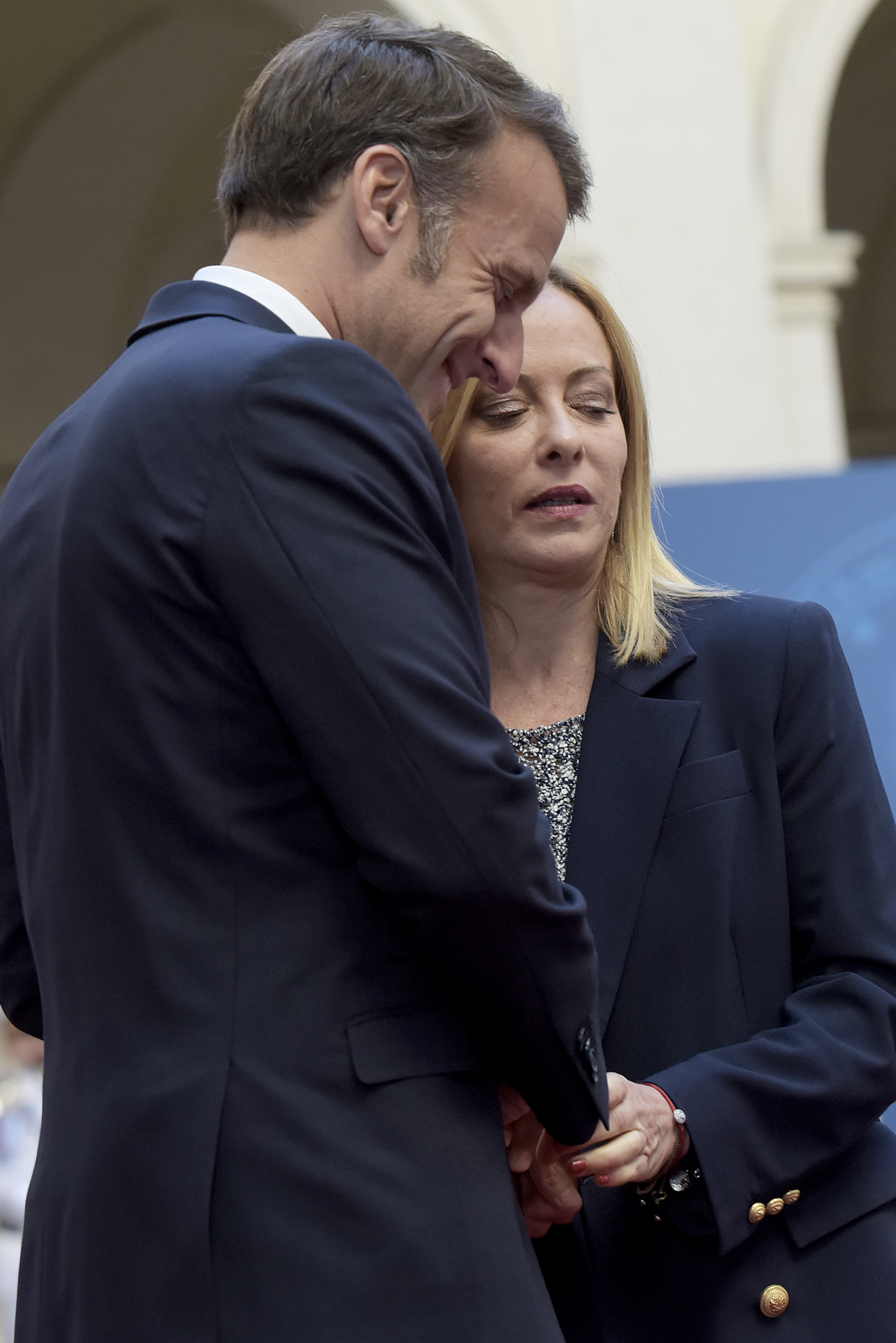 Emmanuel Macron appears to speak softly near Giorgia Meloni’s ear while she closes her eyes, maintaining a composed posture. Their hands remain clasped as they stand close.