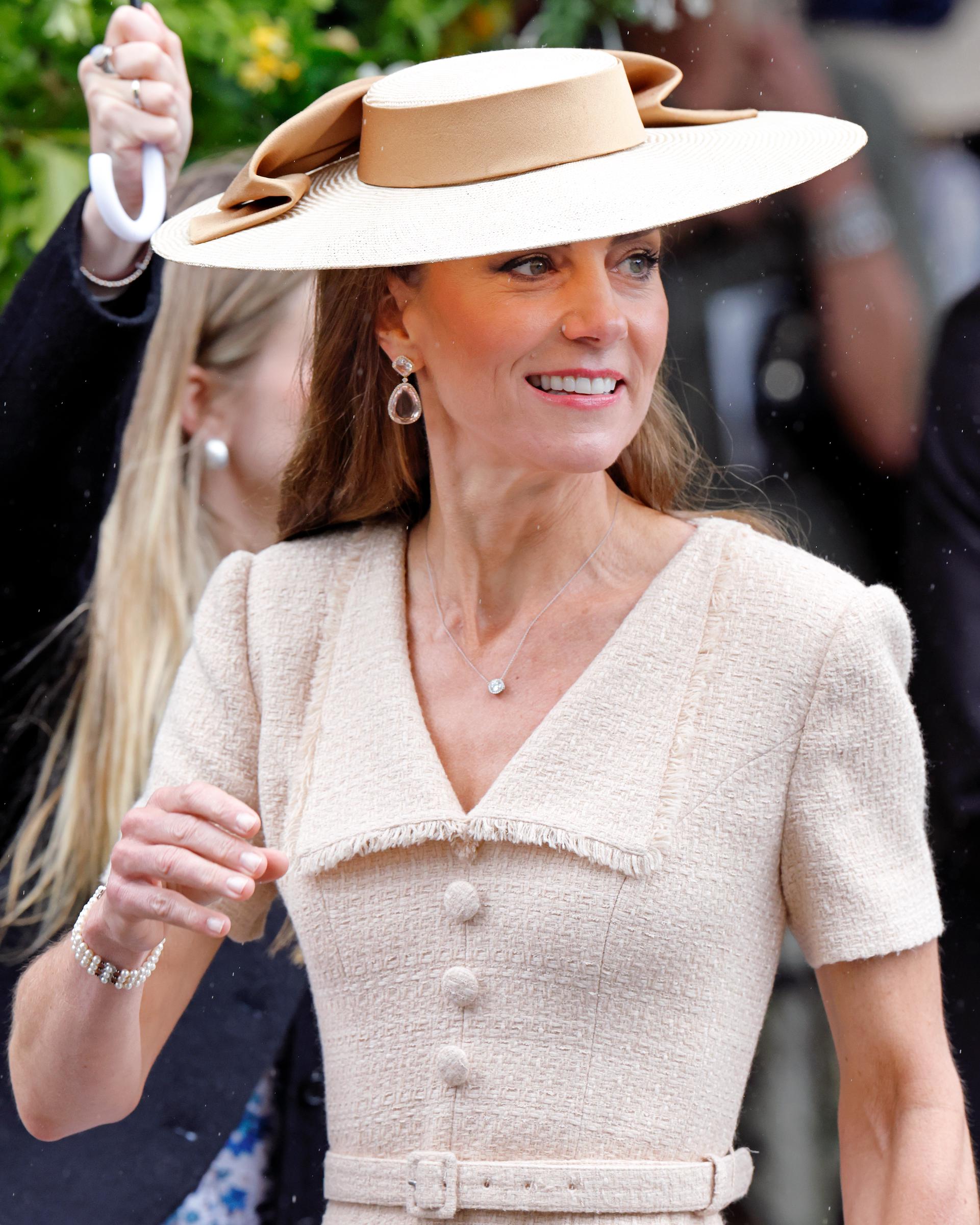 A close-up photo highlights Catherine’s textured cream dress, statement hat with an oversized bow, and elegant jewelry as she arrives at the royal wedding in rainy conditions. | Source: Getty Images