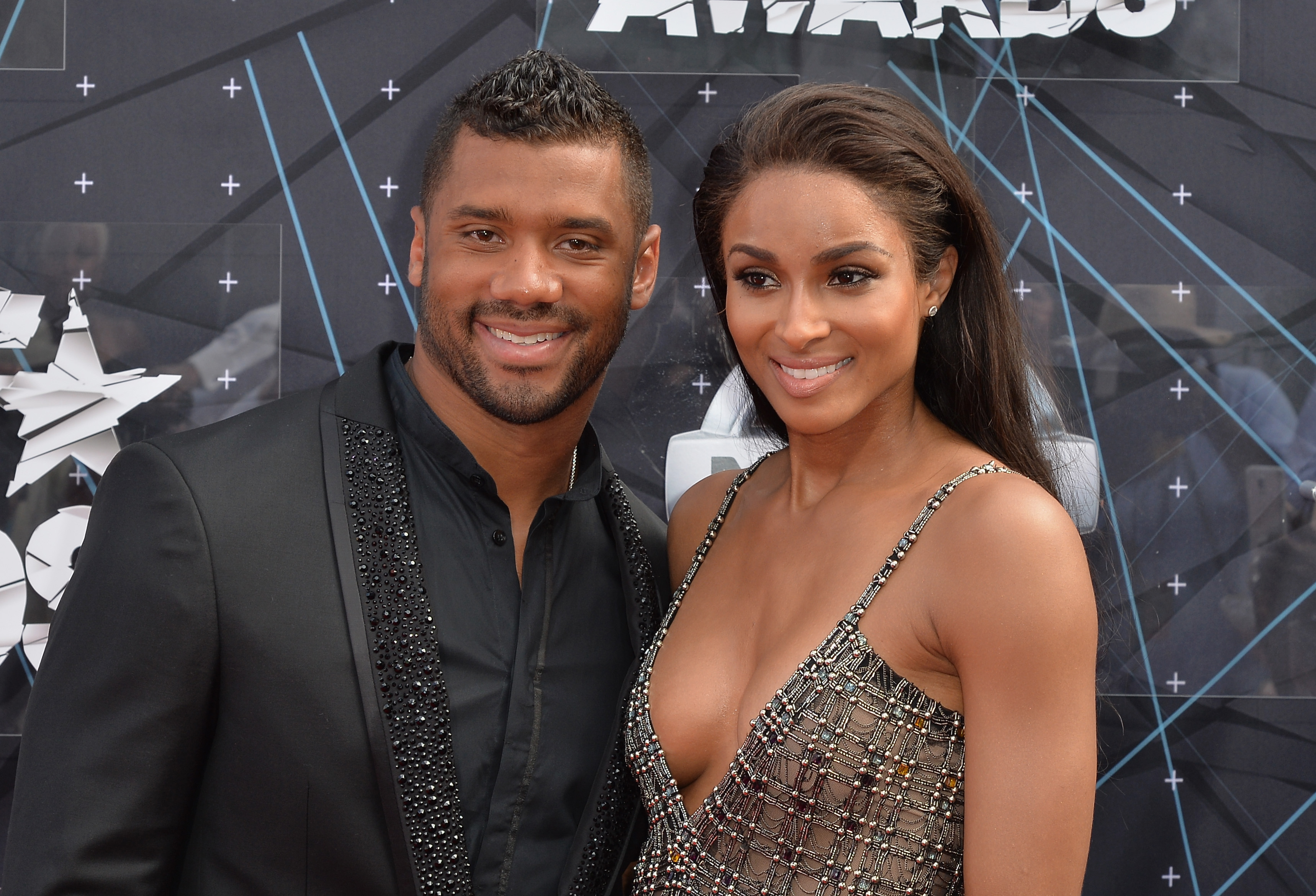 Russell Wilson and Ciara pose on the red carpet in coordinated black looks, standing closely together with an easy, natural chemistry between them. Both wear warm smiles, leaning slightly toward each other as they present a polished yet relaxed presence.